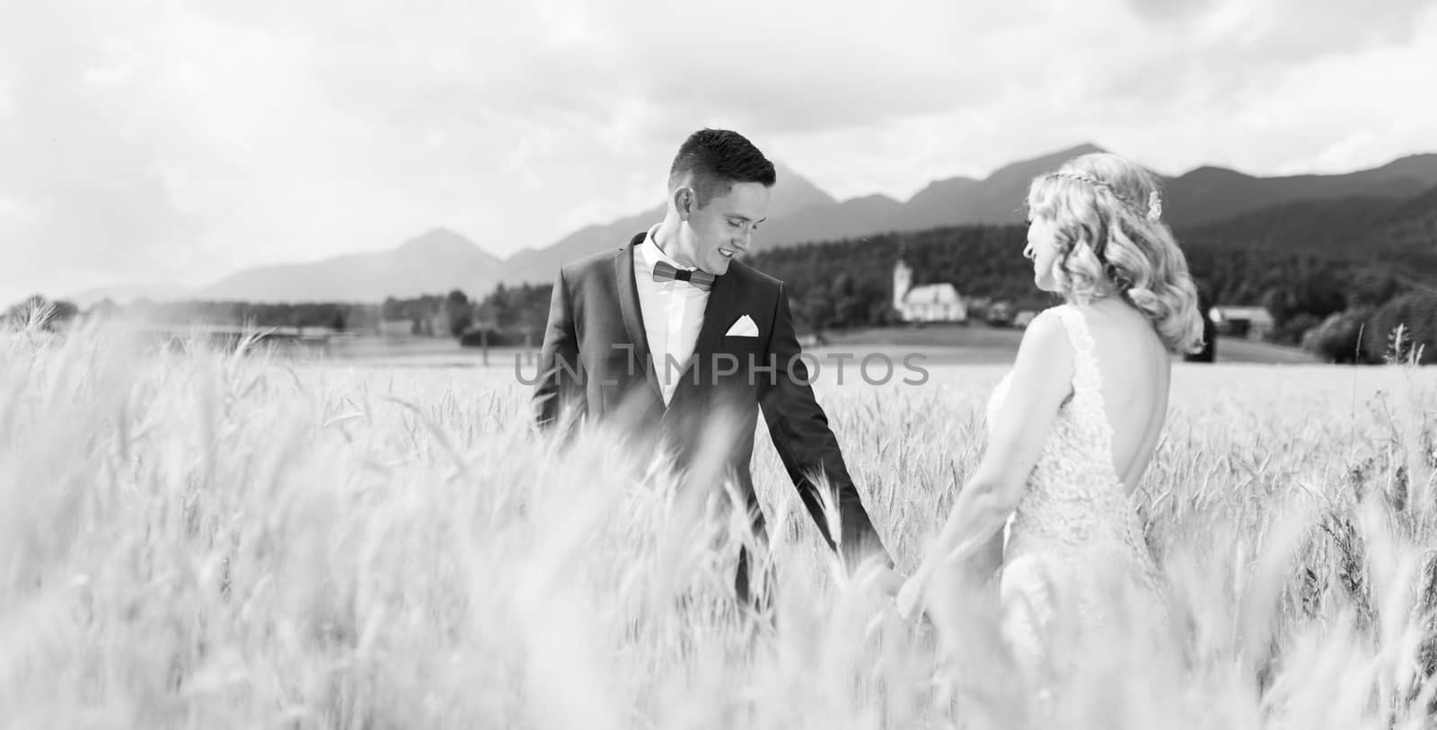 Groom and bride holding hands in wheat field somewhere in Slovenian countryside. Caucasian happy romantic young couple celebrating their marriage. Black nad white photo.