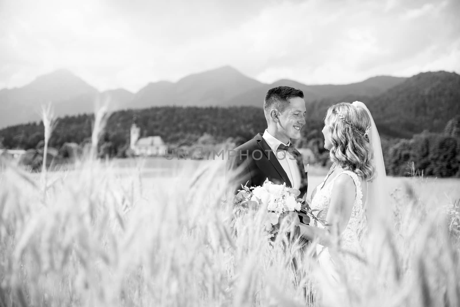 Groom hugs bride tenderly in wheat field somewhere in Slovenian countryside. Caucasian happy romantic young couple celebrating their marriage. Black nad white photo.
