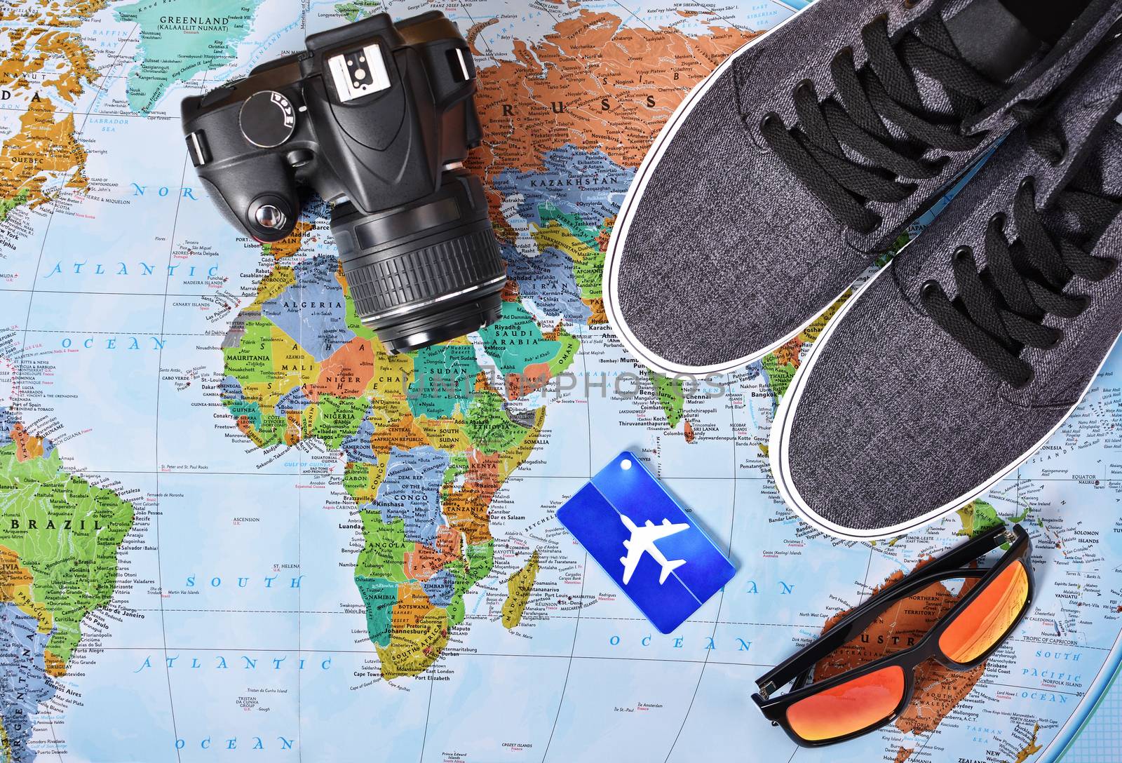 Top view of camera, airplane luggage tag, sunglasses and sneakers on a map of the world. These are my travel essentials for a trip around the world or just a weekend getaway.