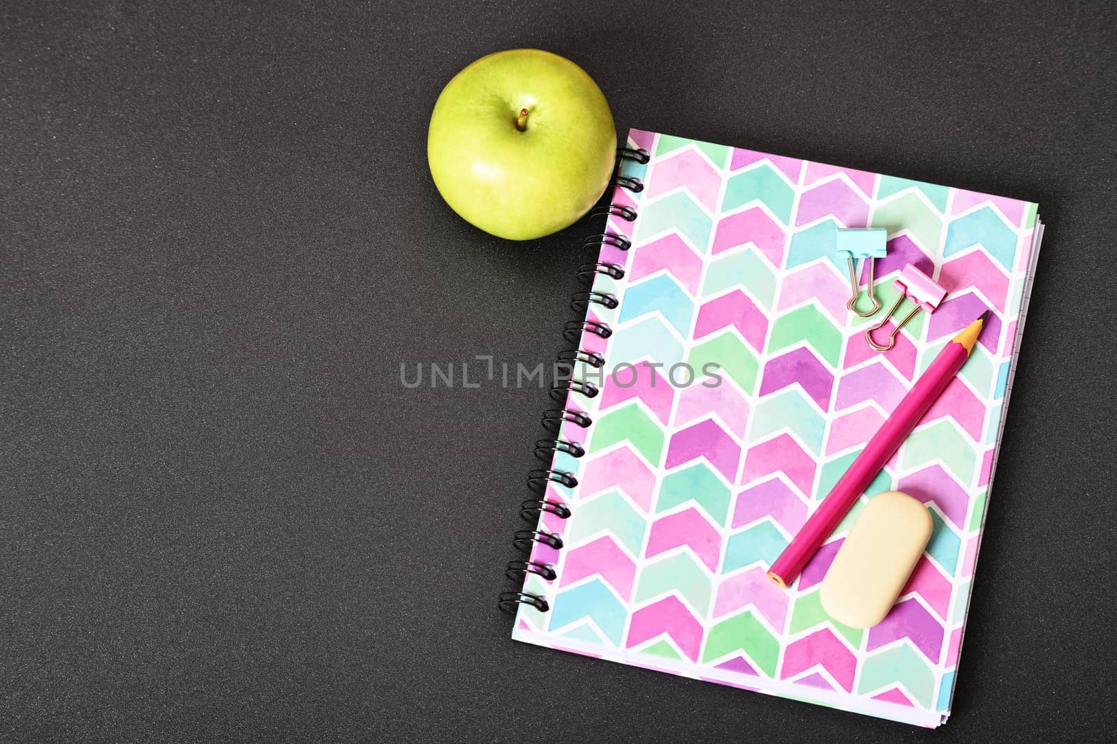 Top view of girly pink notebook with pencil, eraser, binder clips and apple on black background.