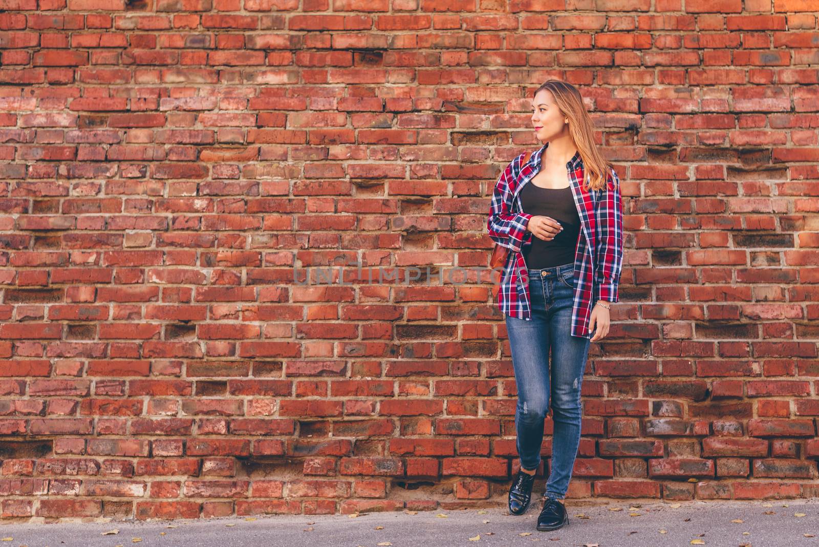 Portrait of young woman in checkered shirt and blue jeans standing against red brick wall