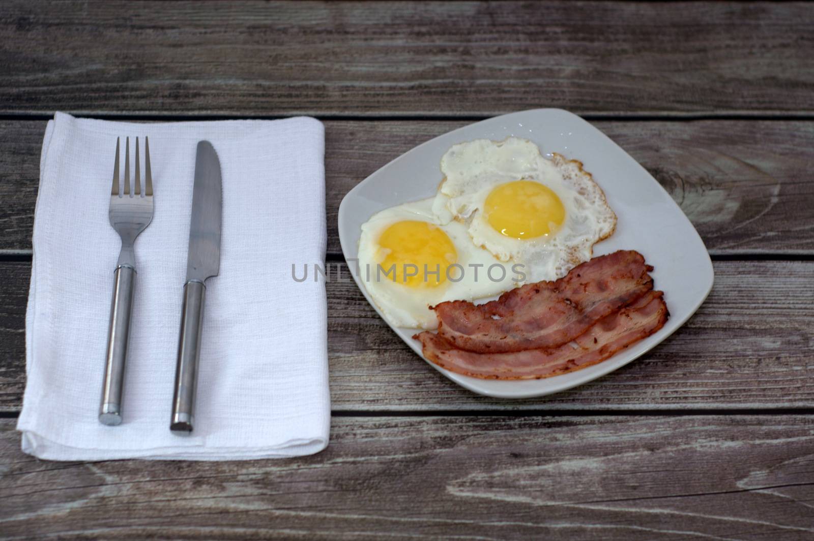 A plate with fried eggs and bacon on the table, next to a napkin with cutlery.