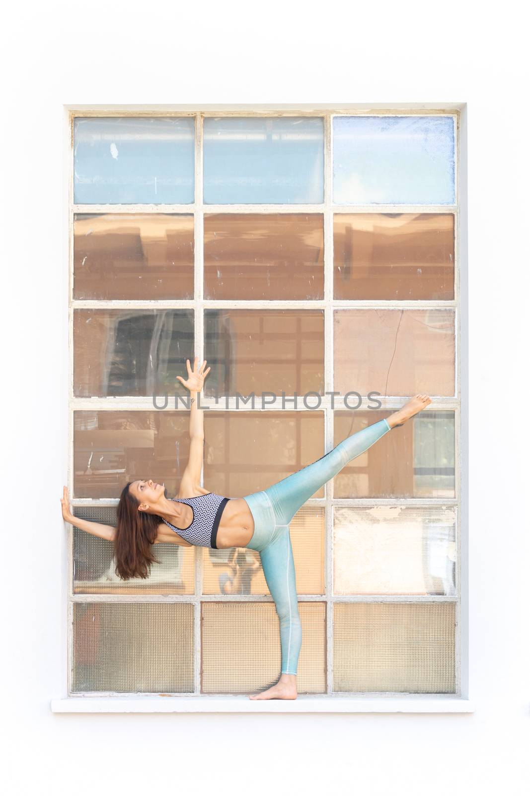 Fit sporty active girl in fashion sportswear doing yoga fitness exercise in front of big industrial window frame. colorful reflections in window glass. Outdoor sports, urban style yoga.