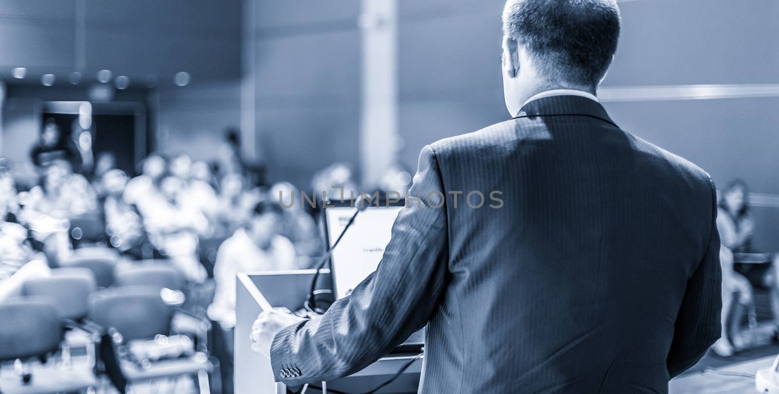 Speaker giving a talk on corporate business conference. Unrecognizable people in audience at conference hall. Business and Entrepreneurship event. Blue toned grayscale image.