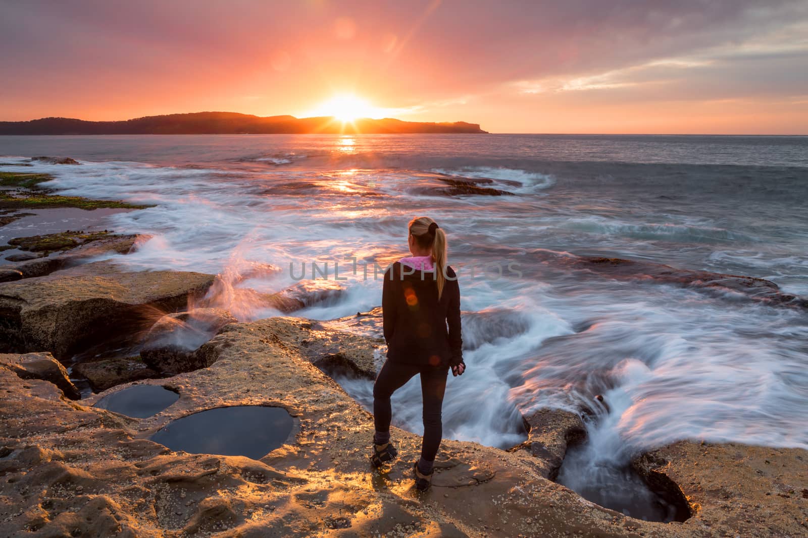 Sunflare kissing the shoulder of a woman standing  by the rocky coastal shore watching the sunrise