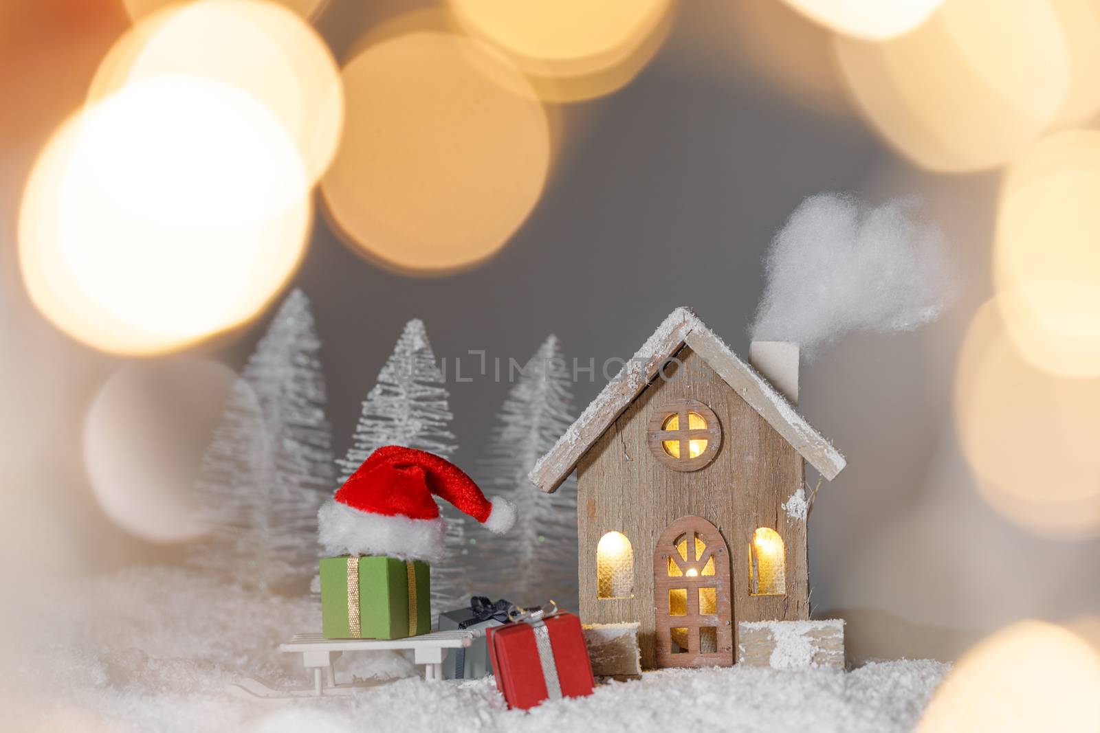 Christmas card with cozy small decorative house , gifts and Santa Claus cap on sleigh on snow over fairy tale winter fir forest background and falling snow . New Year design postcard . Copy space for text