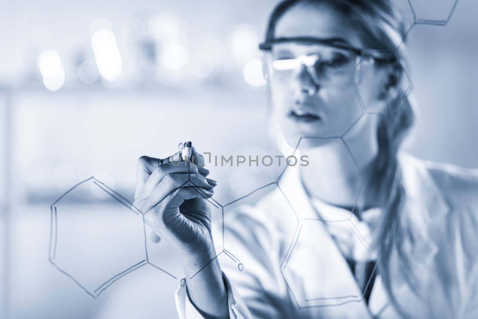 Portrait of a confident female health care expert in life science laboratory writing structural chemical formula on a glass board. Healthcare and modern life science concept. Blue toned image.