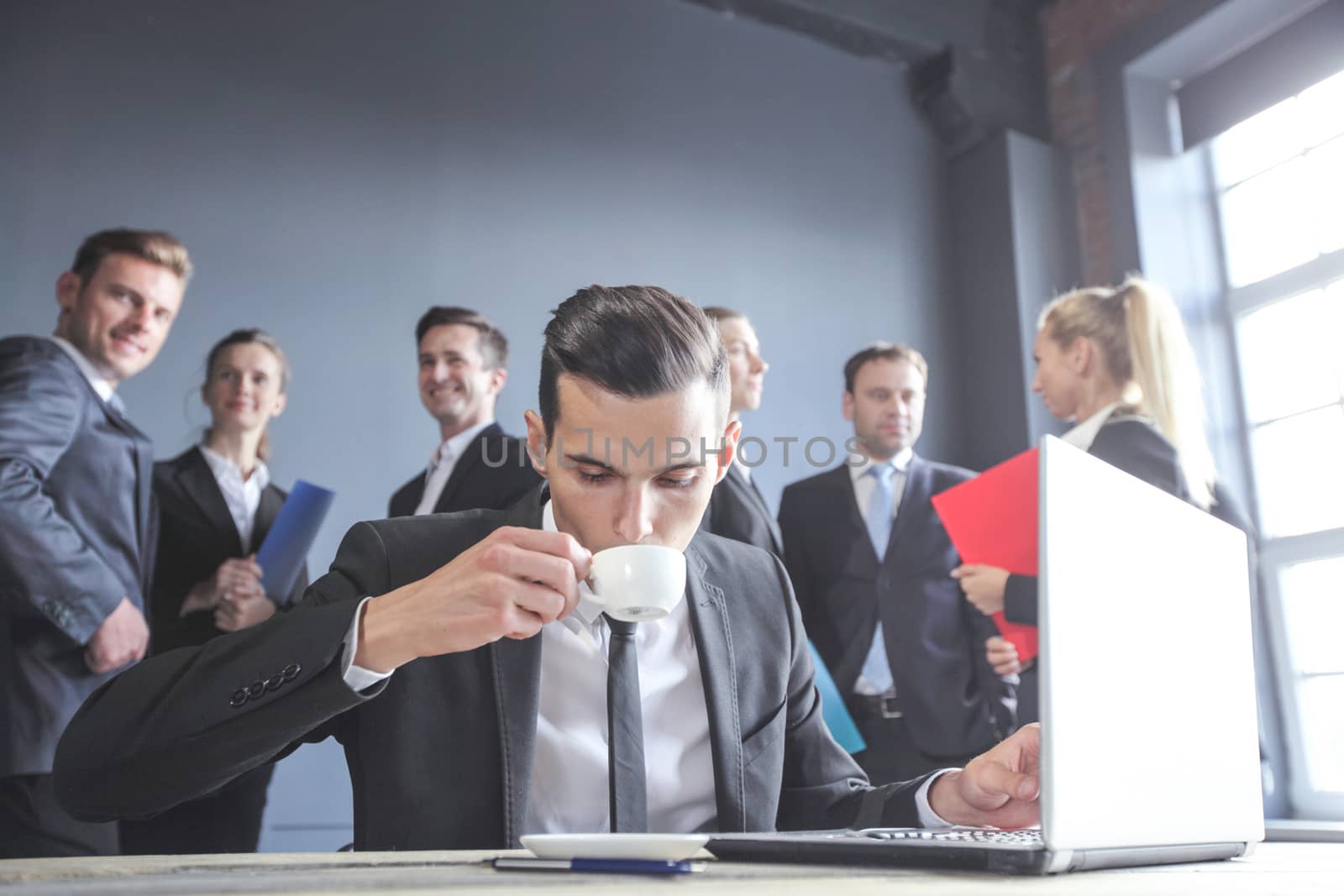 Business man working with laptop and drinkig coffee in office, his team of coworkers on background