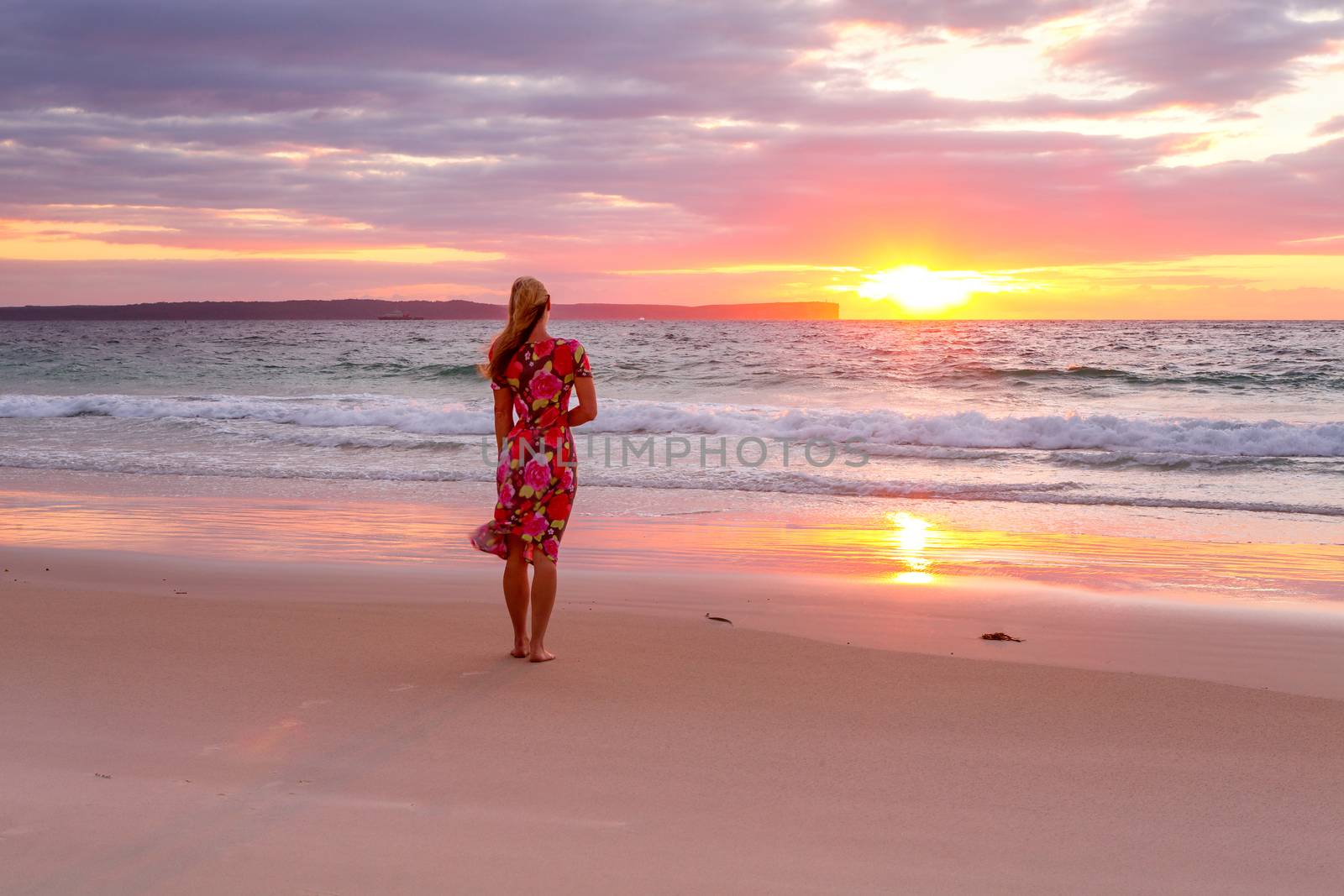 Woman standing on the seashore watching the sunrise.  Its reflection in the wet sand.  She is wearing a beach dress that is flowing in the breeze.