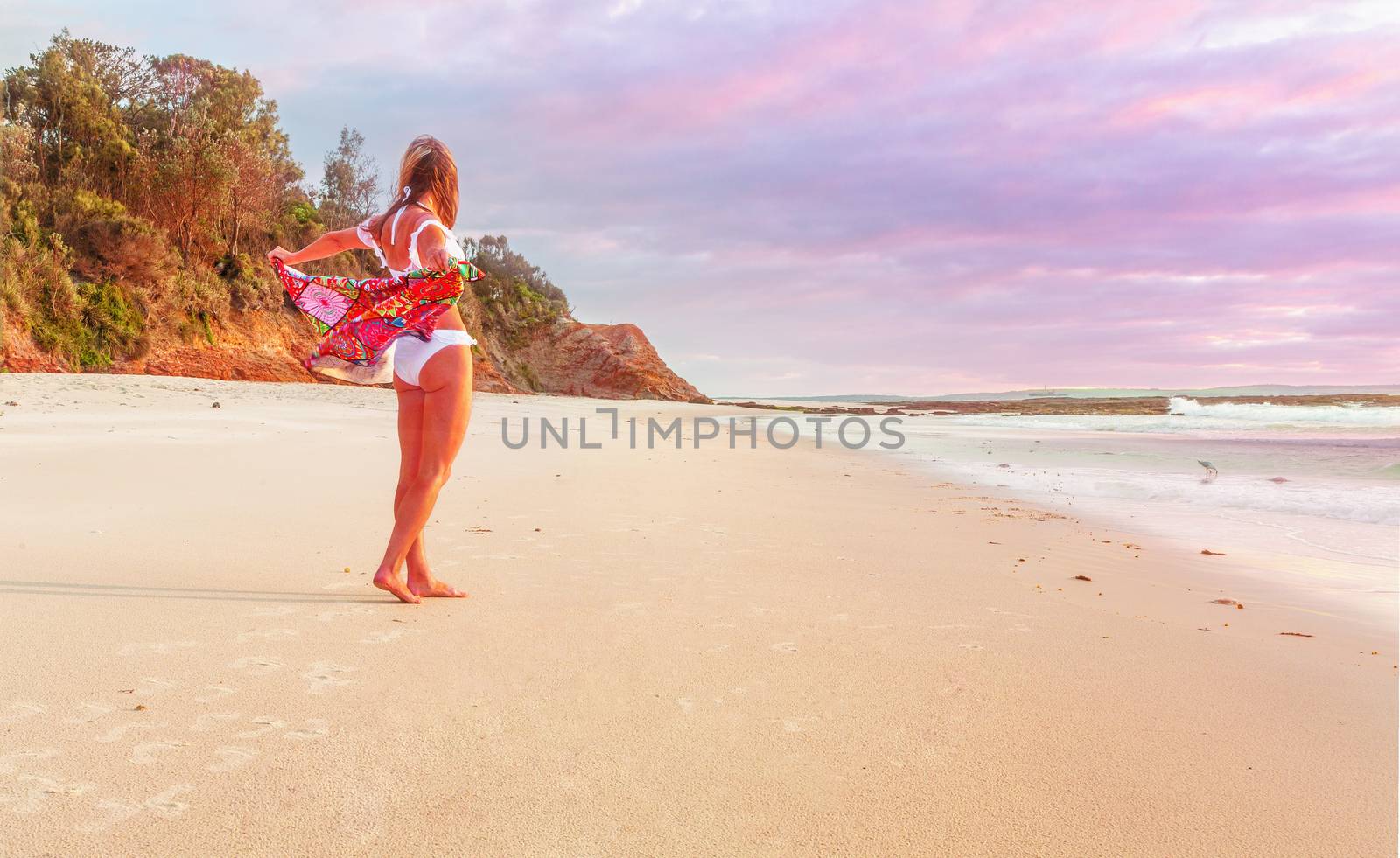 Woman standing on the sandy sea shore with beach towel flapping behind her.  There is some subtle colour in the clouds from the sunrise