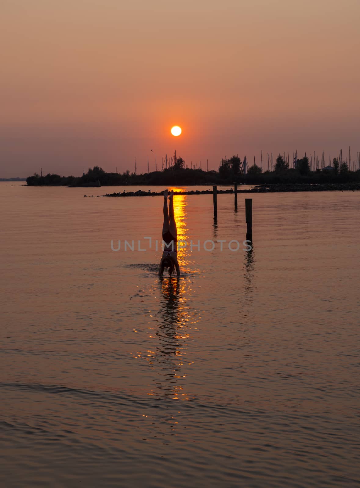 girl doing a handstand in the water during sunset with the harbor of hellevoetsluis and the boats as background