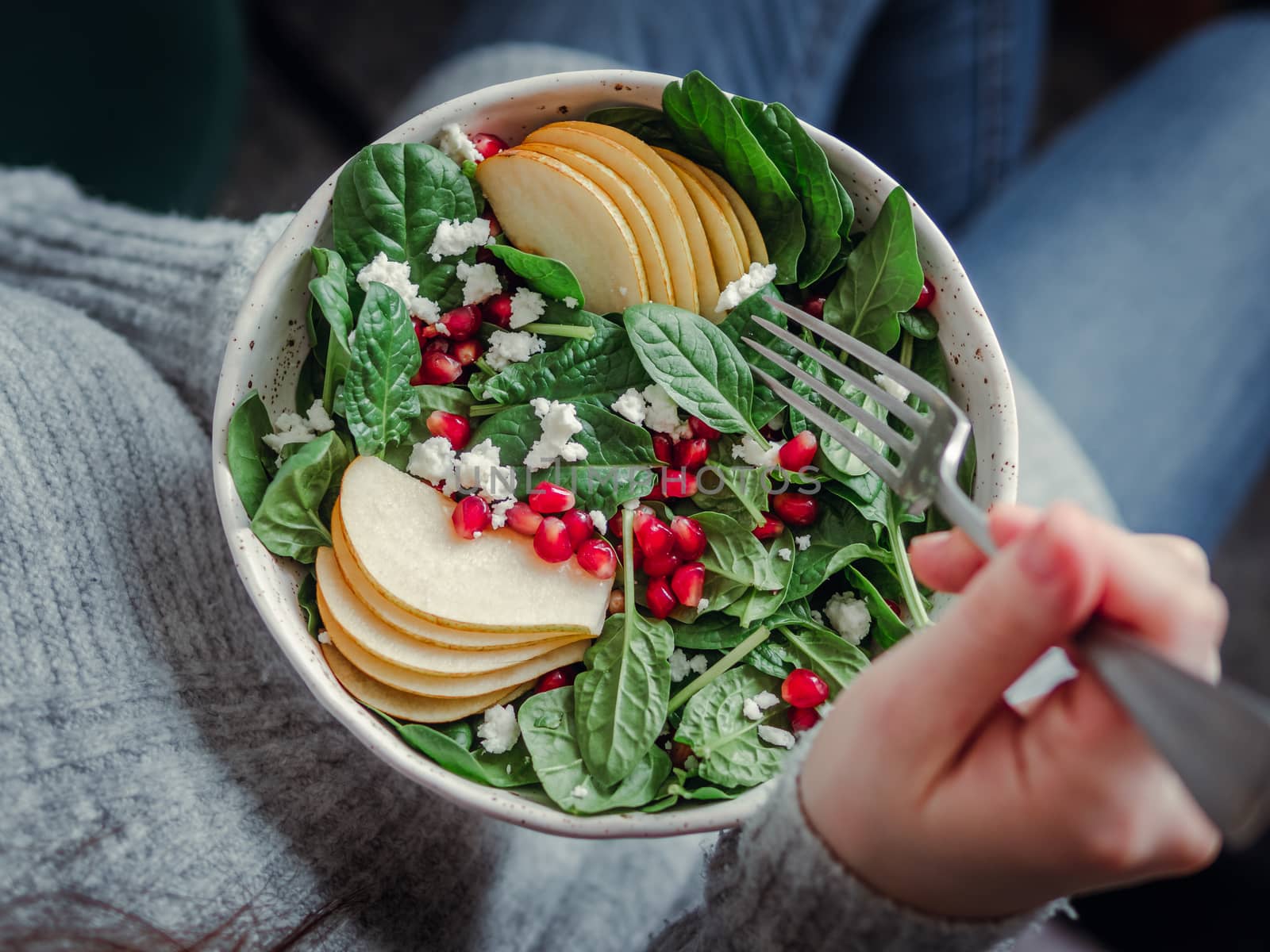 Woman in jeans and sweater holding vegan salad bowl with spinach, pear, pomegranate, cheese. Vegan breakfast, vegetarian food, diet concept. Girl in jeans holding fork with legs, body, hands visible