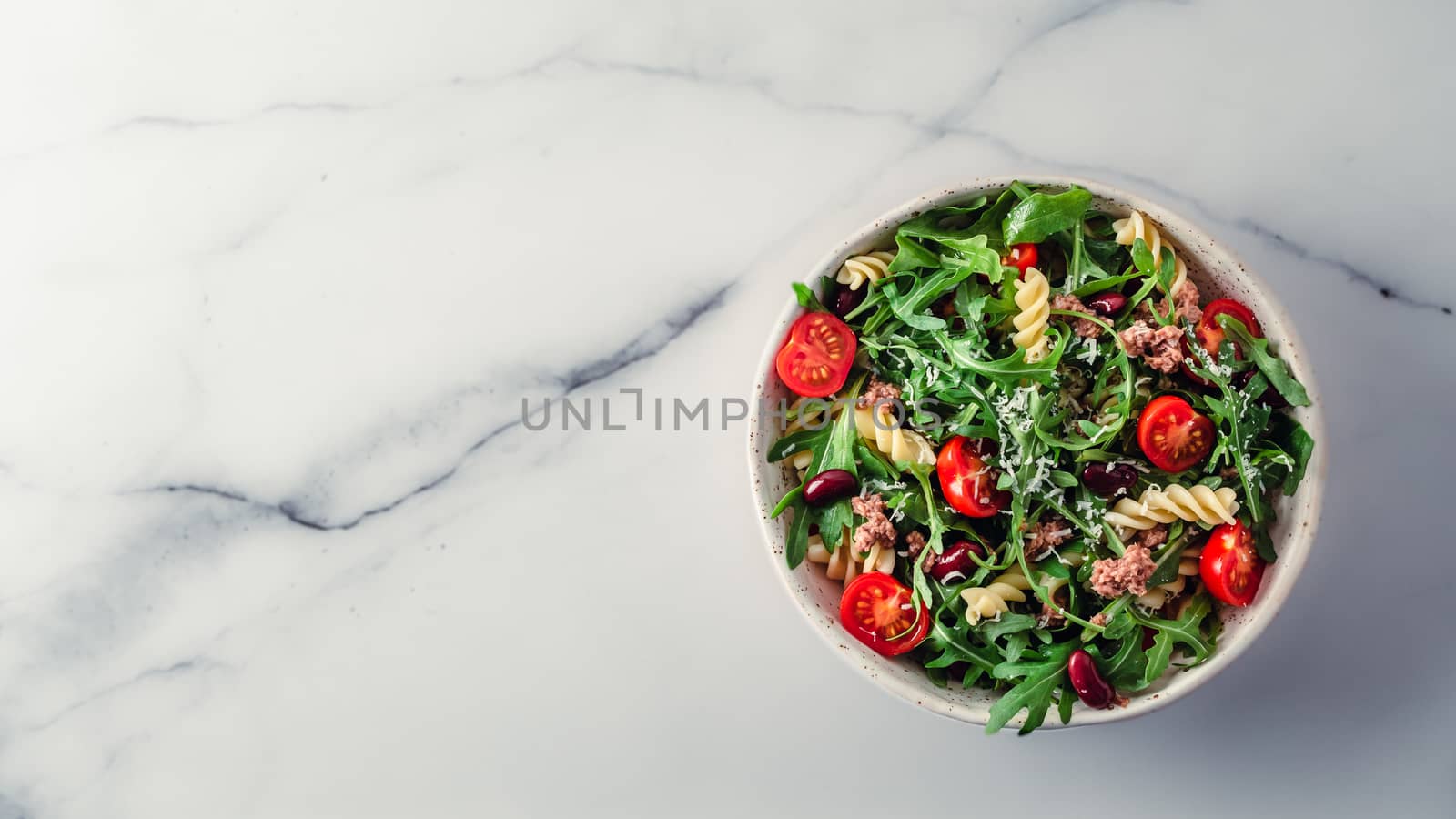 Warm salad with tuna, arugula, tomatoes, red bean, pasta. Idea and recipe for healthy lunch or dinner. Bowl with warm salads on marble tabletop. Healthy dinner. Top view. Copy space for text. Banner