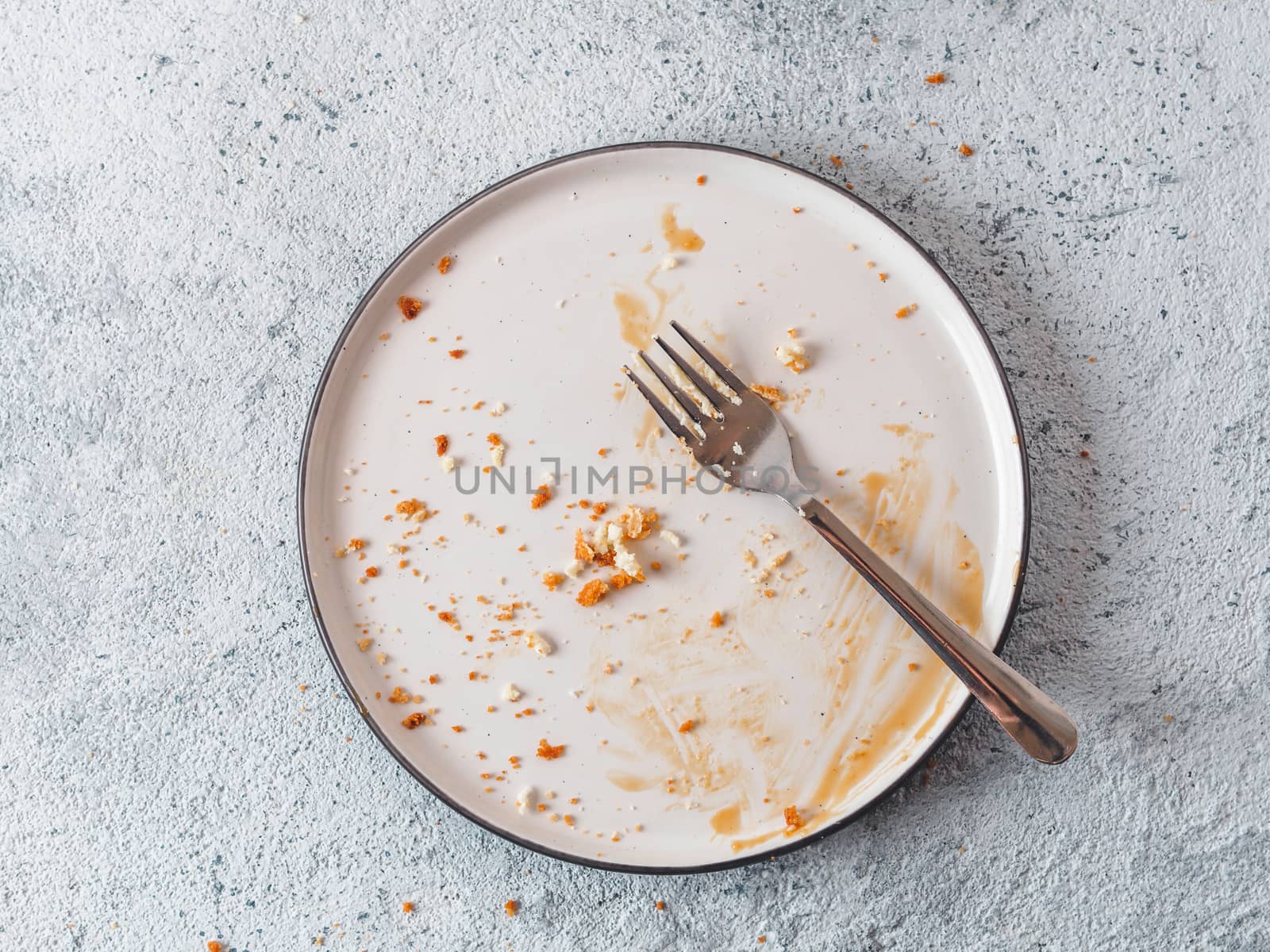 Empty dirty dish after cheesecake with caramel sauce and dessert fork. White rustic trendy modern fashionable dirty plate on gray cement background, top view or flat lay. Copy space for text