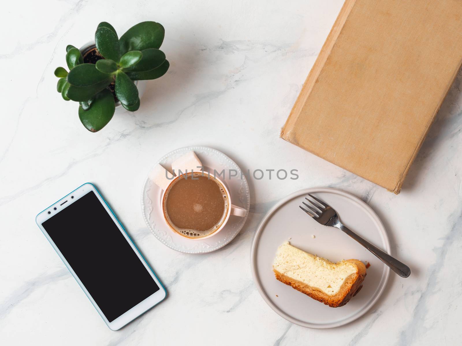 Coffee and cheescake on white marble tabletop. Top view or flat lay. Coffee cup, piece of cheesecake, smartphone, book and succulent. Home office, female workspace concept