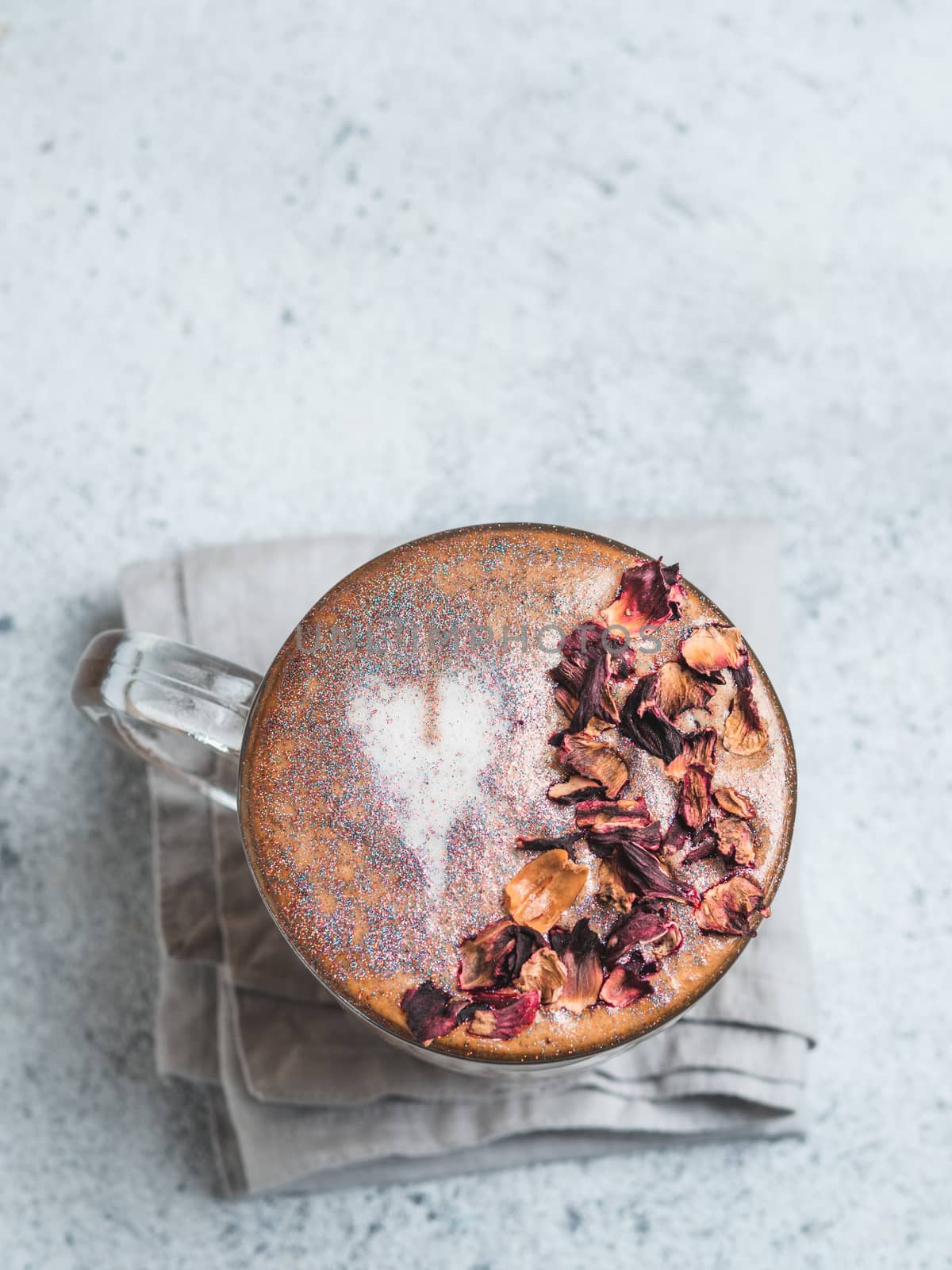 Trendy Coffee with edible glitter and dried rose petals. Cup of sparkly coffee or diamond cappuccino on gray table. Copy space for text. Top view or flat lay. Vertical.
