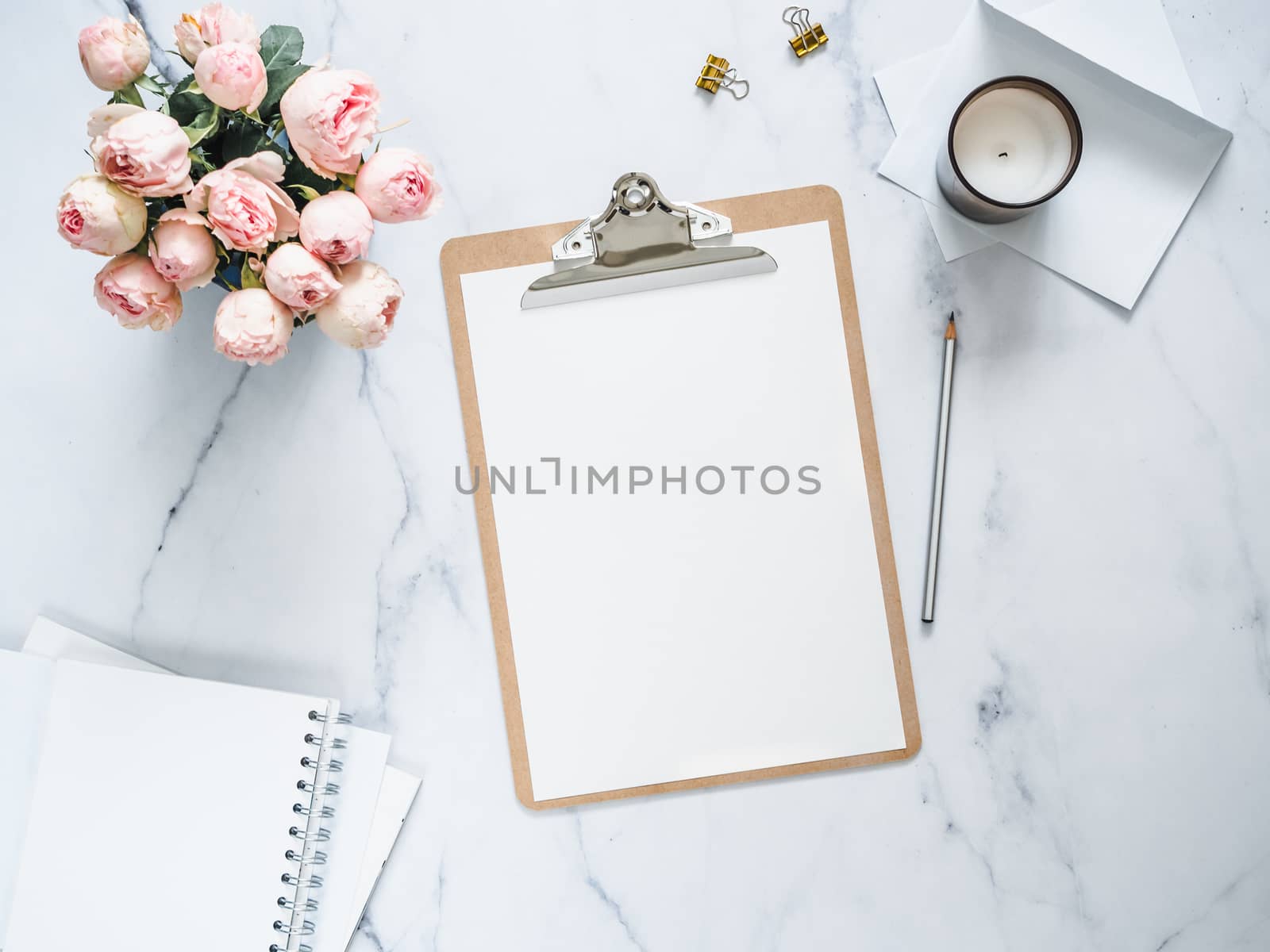 Top view of clipboard with white empty page. Clipboard, flowers, scented candle on white marble. Feminine home office mock up with blank sheet of paper A4 portrait format,copy space for text. Flat lay