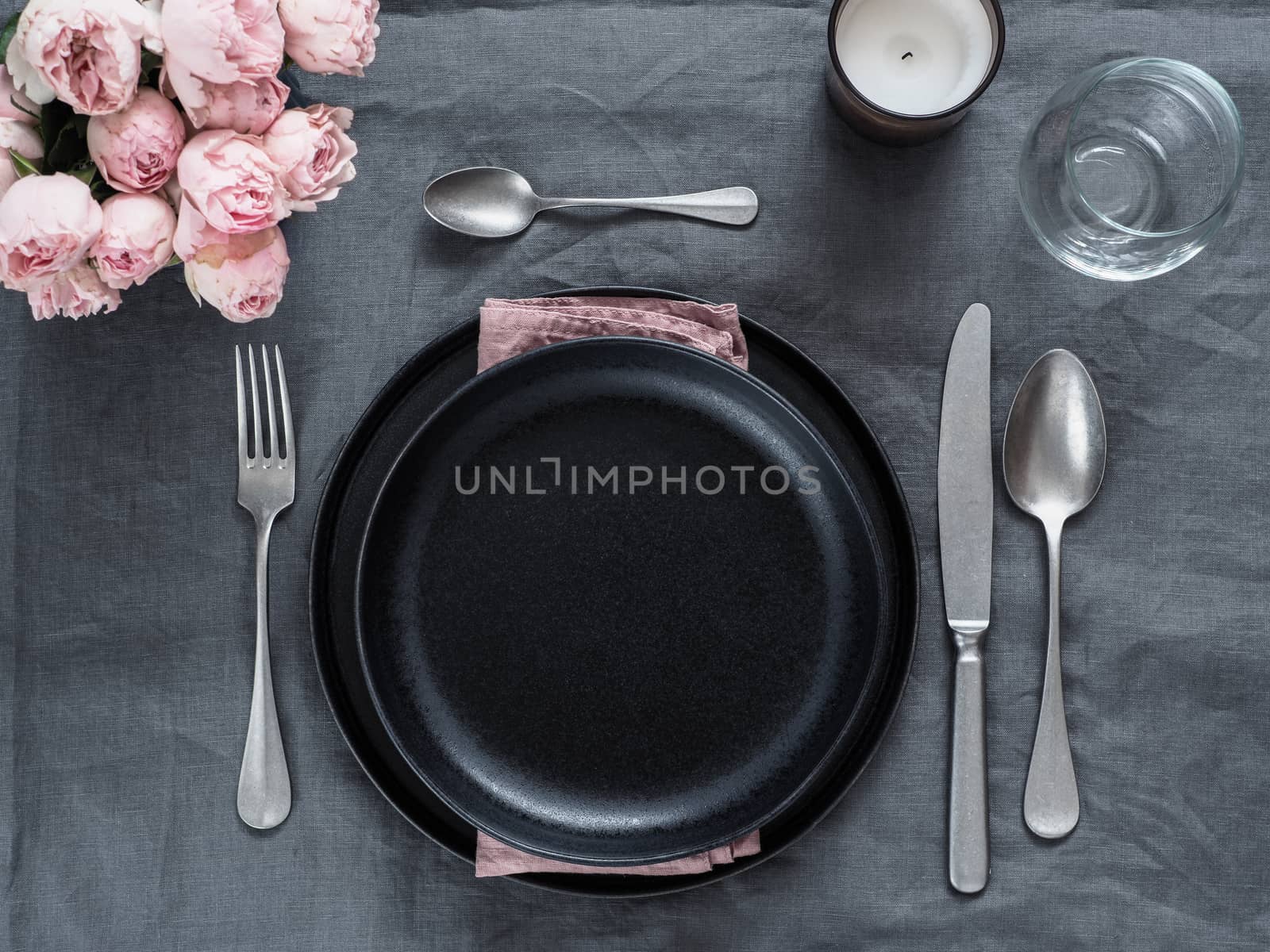 Beautiful table setting on gray linen tablecloth. Festive table setting for wedding dinner with pink spray roses and scented cabdle. Holiday dinner with black plates. Dinner, utensil. Copy space