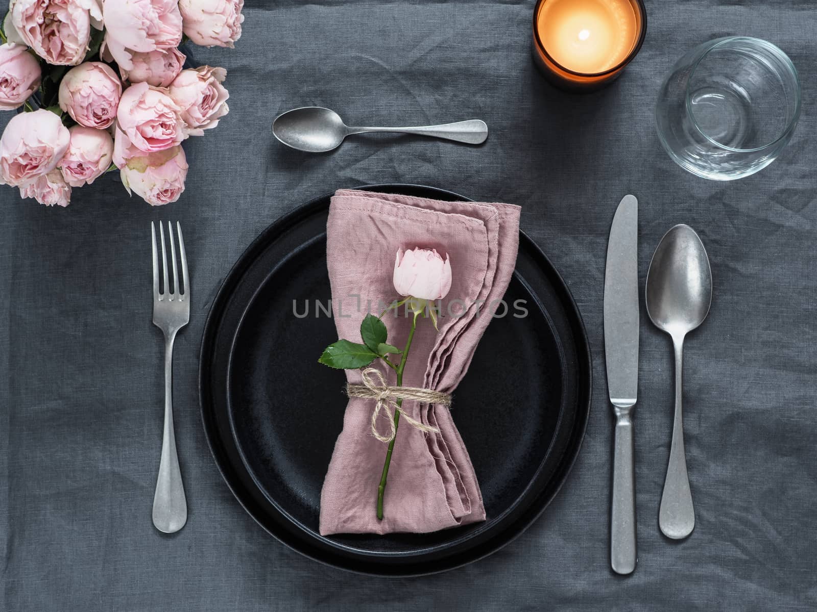 Beautiful table setting with candle on gray linen tablecloth. Festive table setting for wedding dinner with pink spray rose and pink napkin on plate. Holiday dinner with modern craft black plates