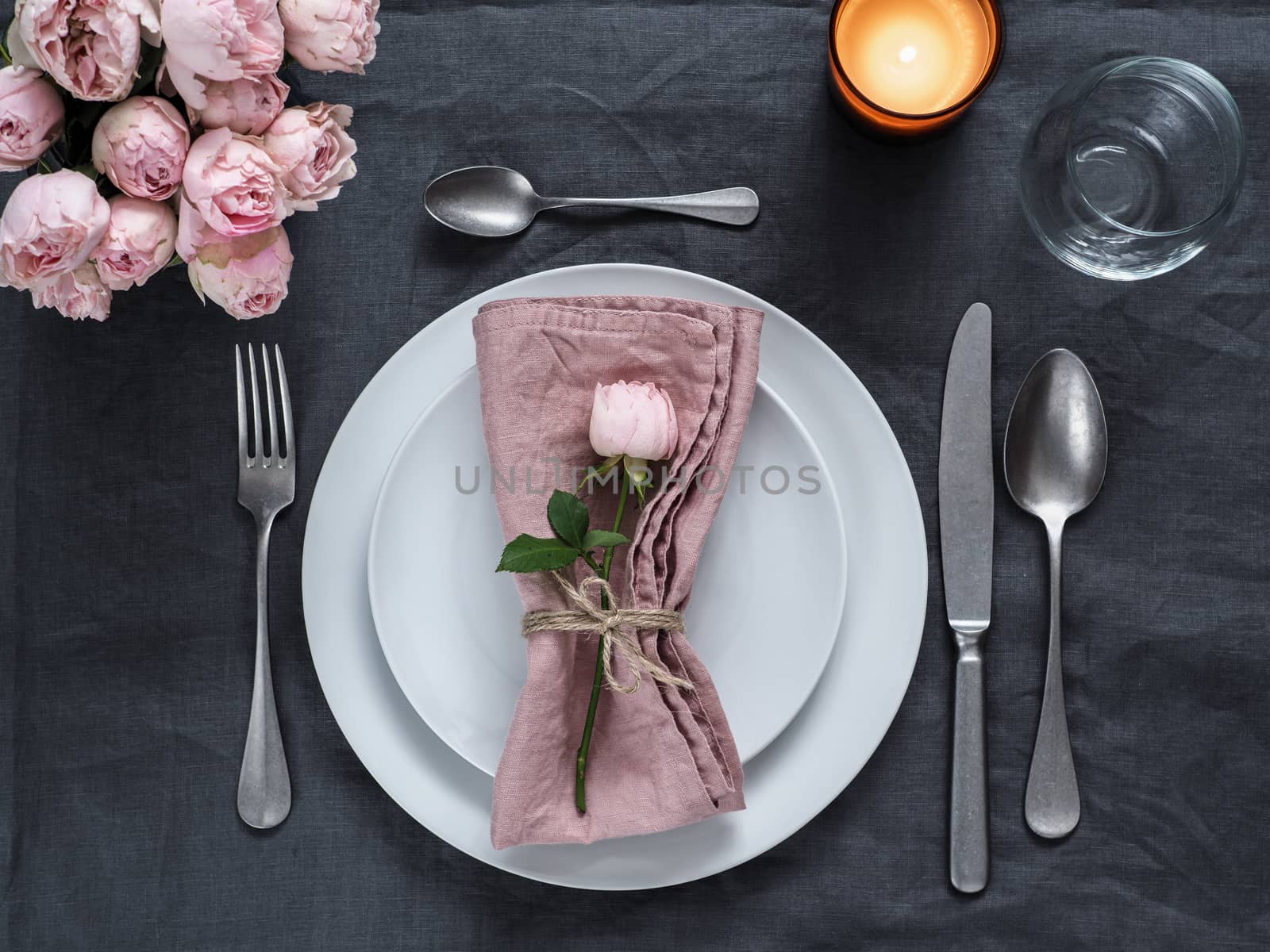 Beautiful table setting with candle on gray linen tablecloth. Festive table setting for wedding dinner with pink spray rose and pink napkin on plate. Holiday dinner with white plates