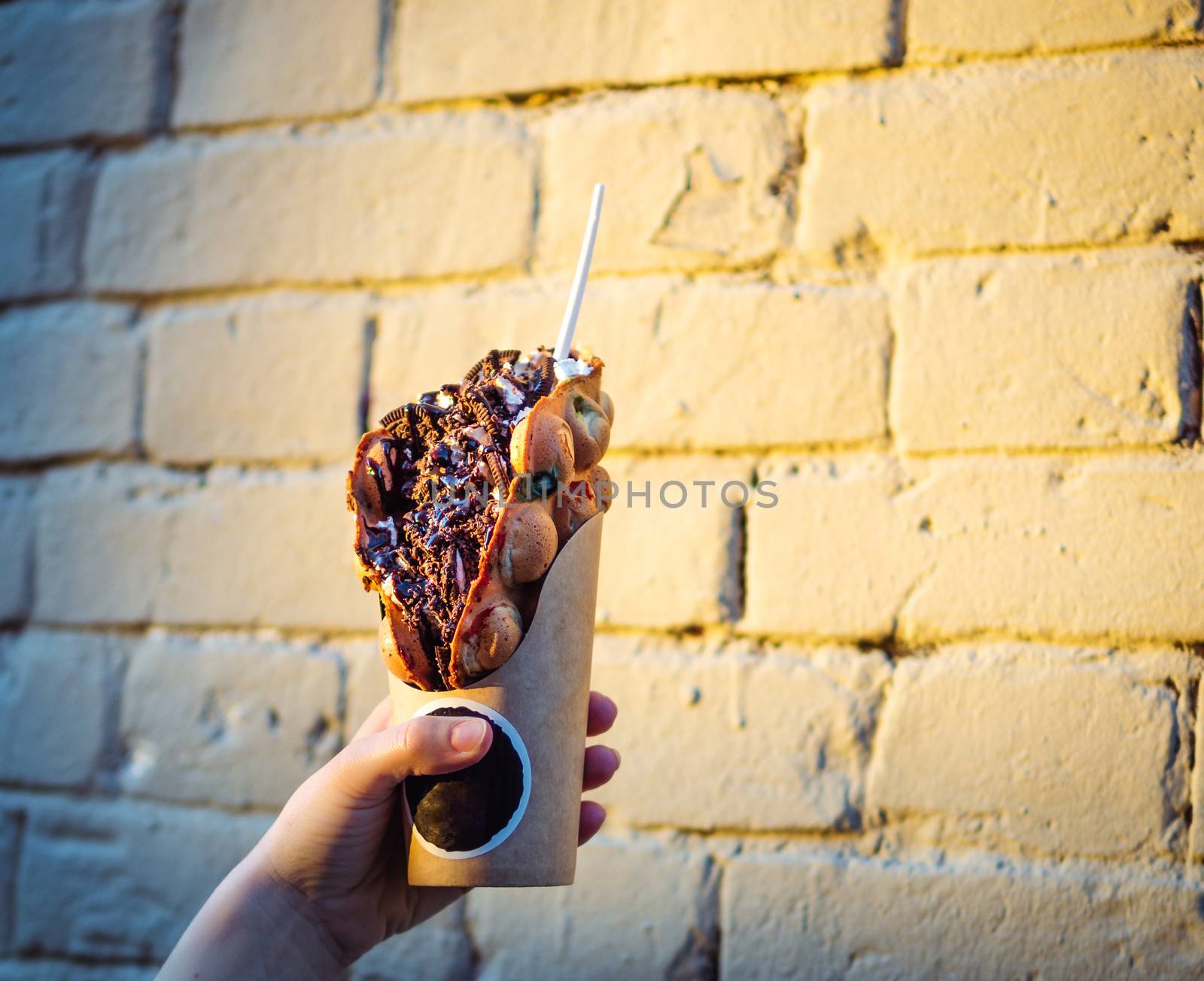 Hong Kong waffles in female hand on brick wall background. Delicious bubble waffle with chocolate biscuit cookies and chocolate syrup in beautiful sunset light. Copy space for text.