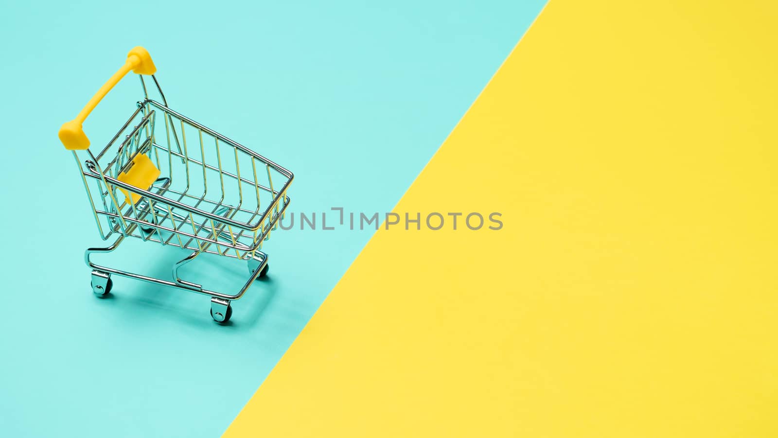 Empty miniature shopping cart on blue and yellow background. Toy trolley on bright colorful background, copy space for text or design.