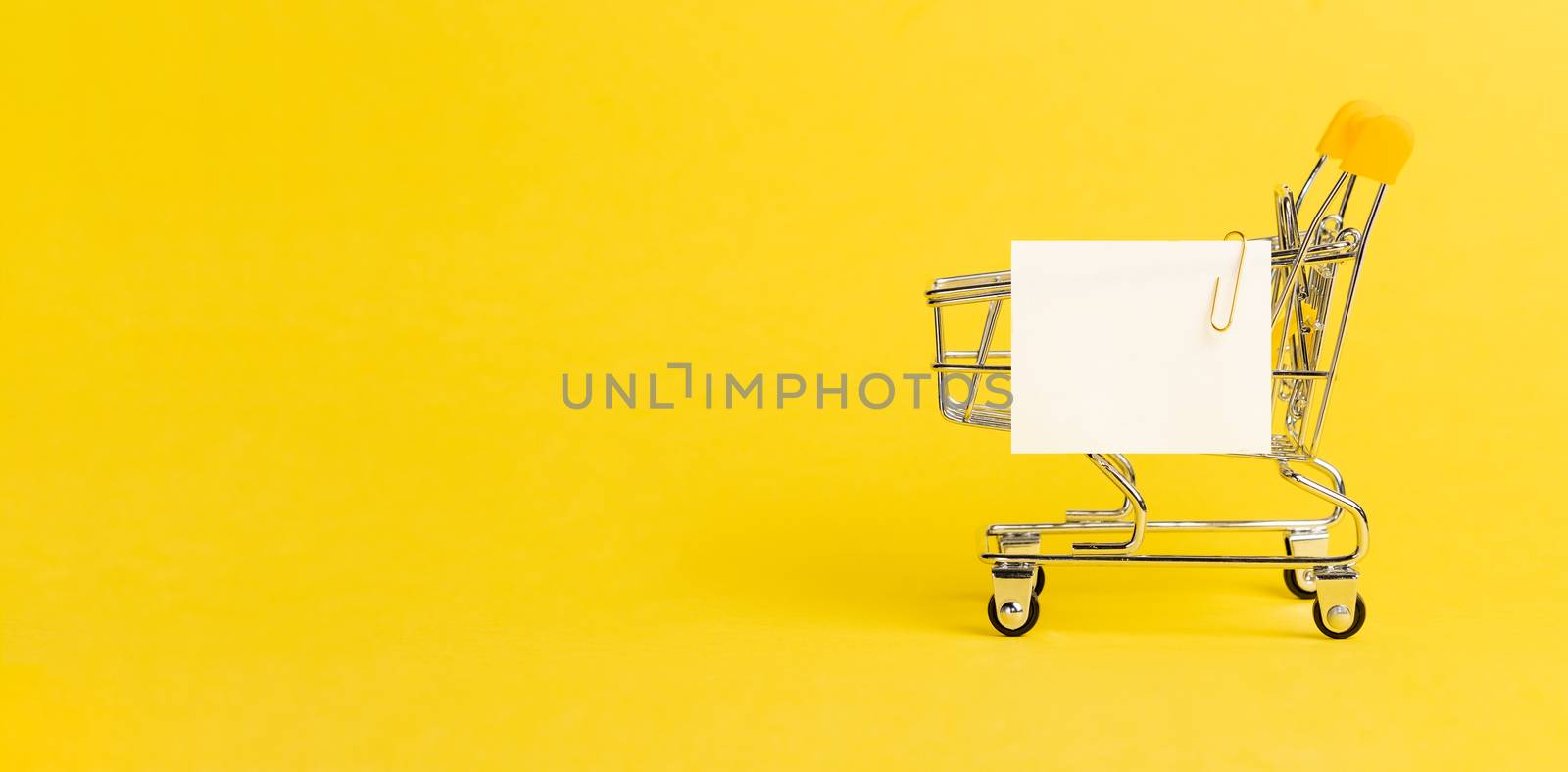 Shopping cart and white paper note list over yellow background. Shopping concept on bright yellow background. Empty white paper note over shopping cart. Copy space for text or design. Banner
