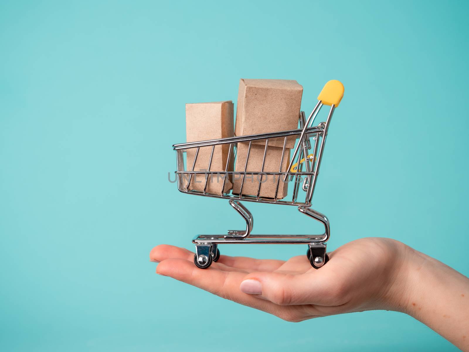 Toy shopping cart with boxes in female hand over blue background. Copy space for text or design. Ssale, discount, shopaholism concept. Consumer society trend