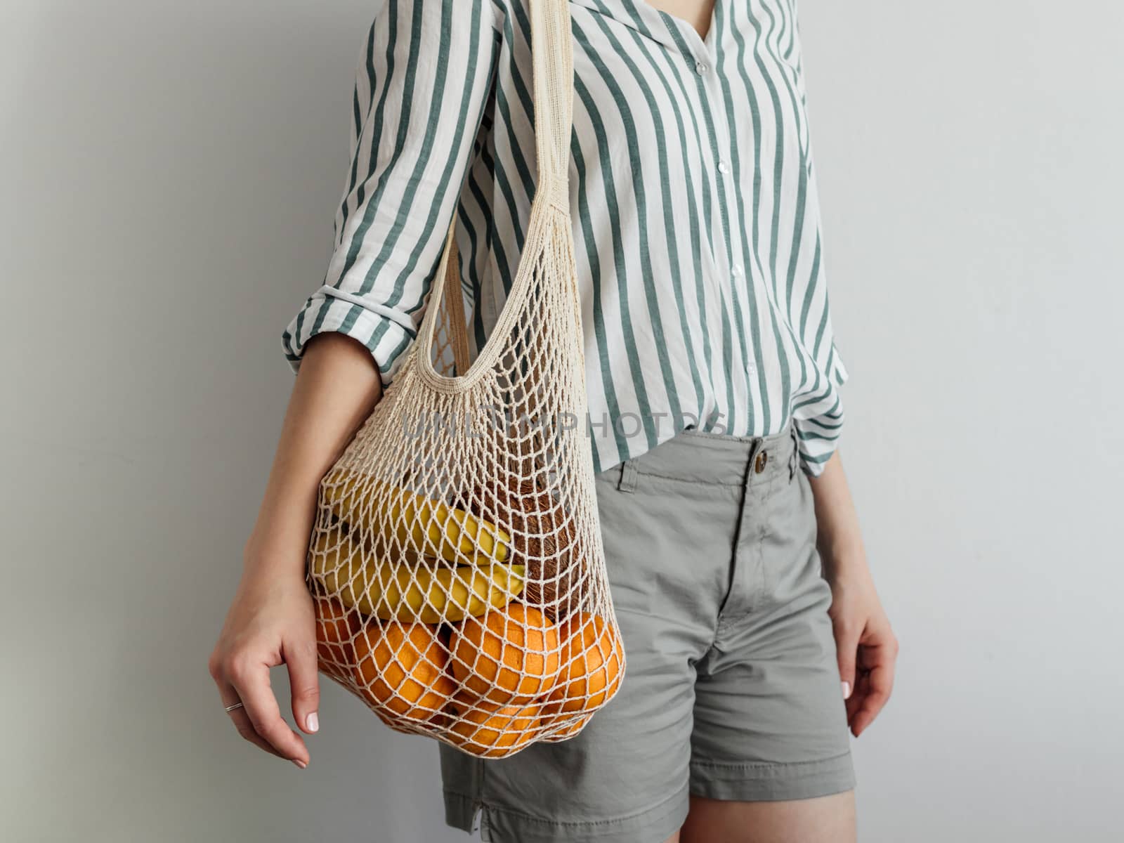 Mesh bag with fruits on female shoulder. Stylish young woman standing with mesh shopping bag on shoulder near light wall. Modern reusable shopping concept.