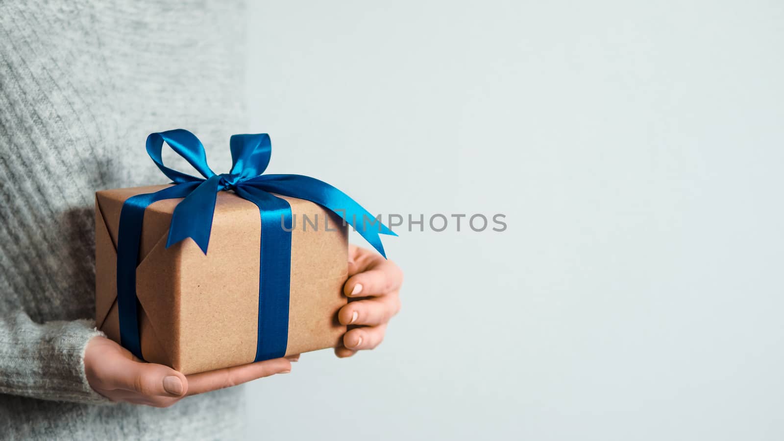 Female hands hold gift box on white background, copy space right. Unrecognizable caucasian girl in gray sweater holding gift box in craft wrapping paper with satin ribbon. Christmas, New year banner
