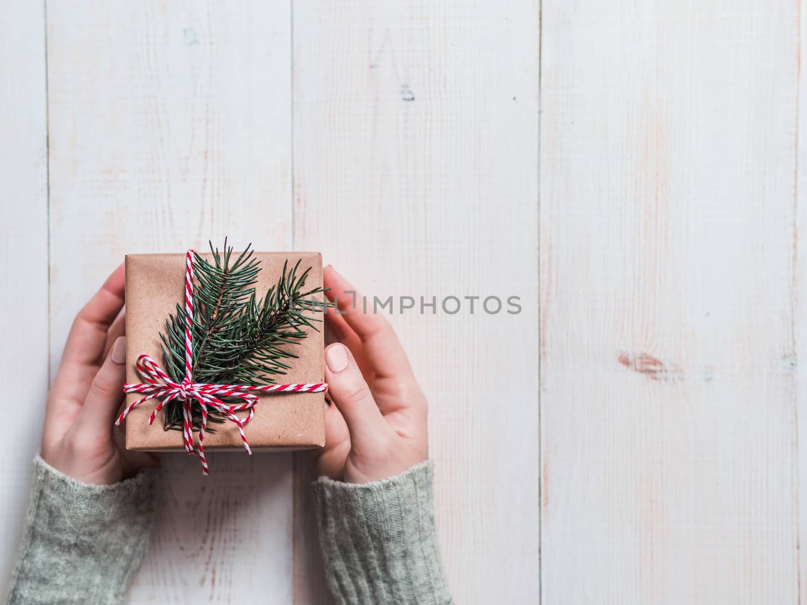 Female hands hold gift box over white wooden background, copy space right. Caucasian girl hands in gray sweater holding gift box in craft wrapping paper with red Christmas string and fir branches