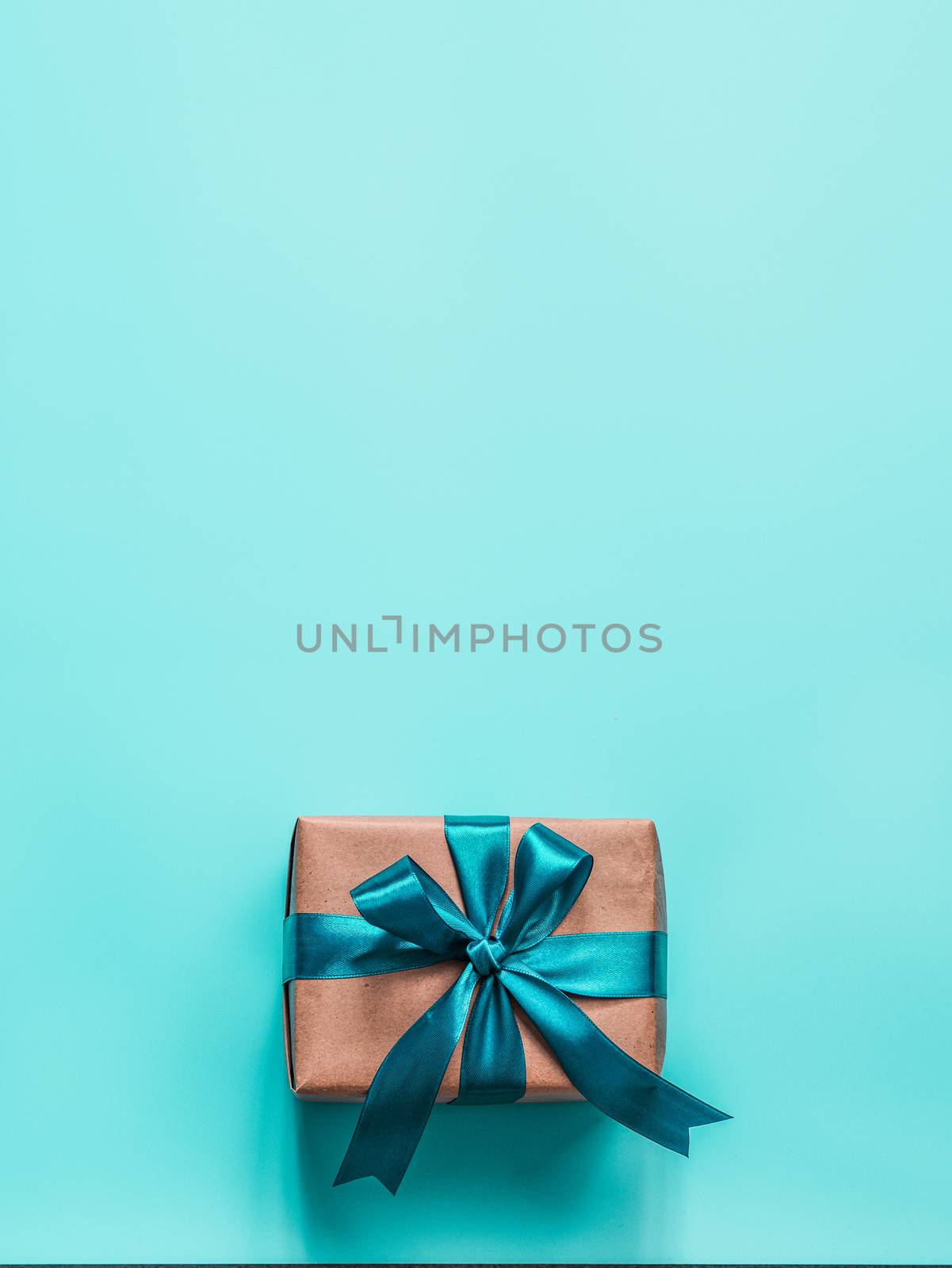 Gift box in craft wrapping paper and green satin ribbon on turquoise blue background, copy space top. Beautiful Christmas, New Year or Birthday present, flat lay or top view. Vertical