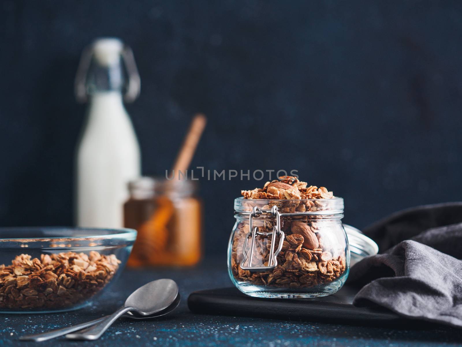 Homemade granola in glass jar on dark table. Ingredients for healthy breakfast - granola, milk and honey. Copy space for text. Low key.