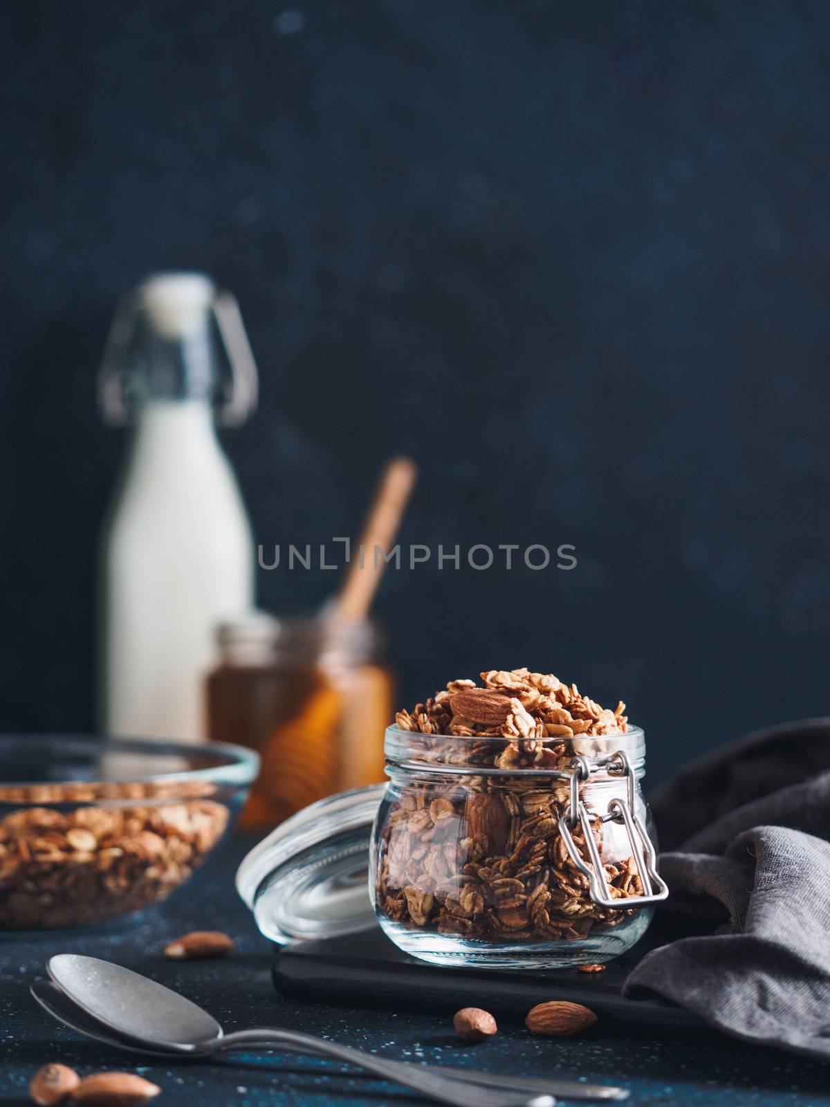 Homemade granola in glass jar on dark table. Ingredients for healthy breakfast - granola, milk and honey. Copy space for text. Low key.