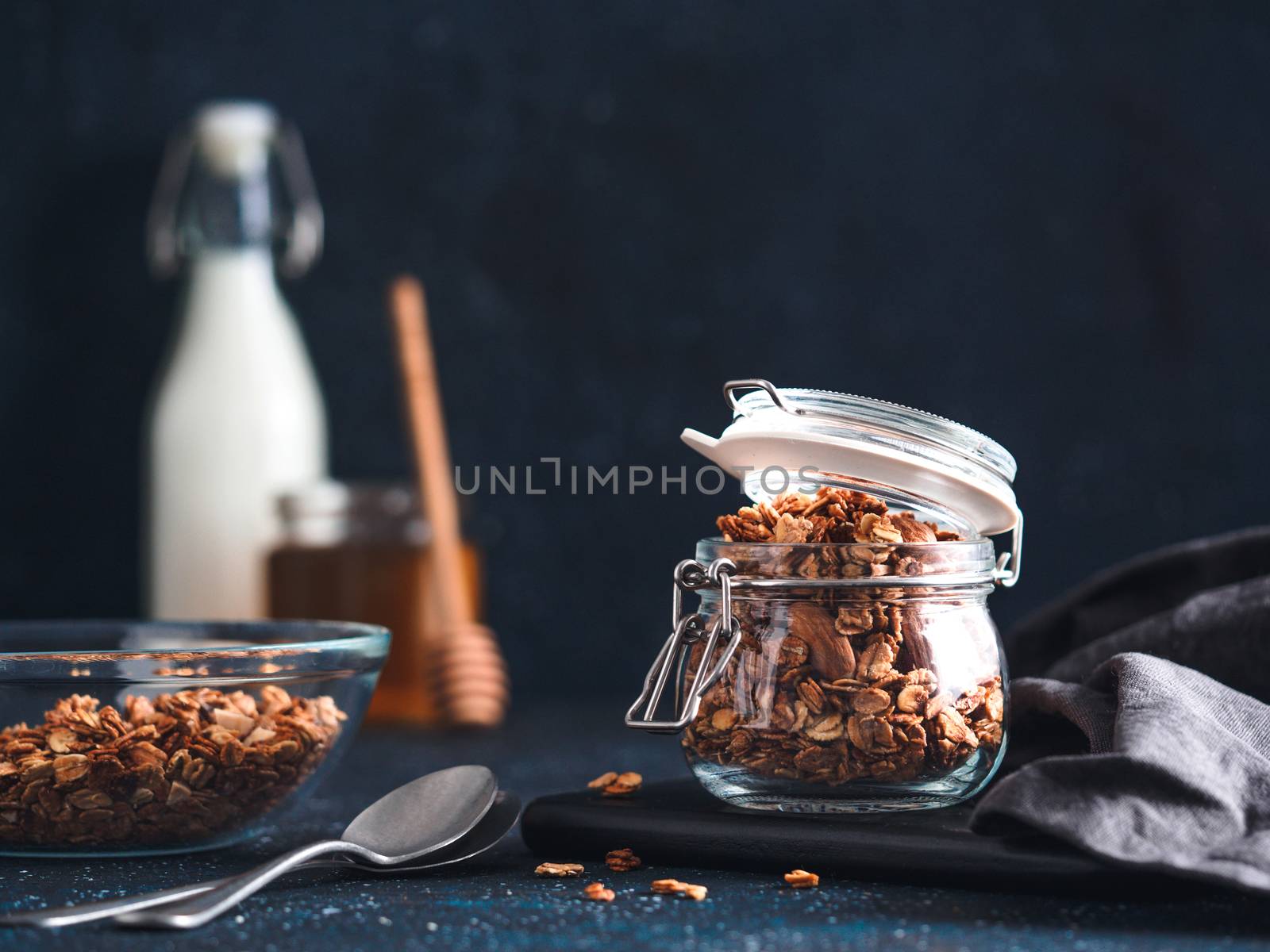 Homemade granola in glass jar on dark table. Ingredients for healthy breakfast - granola, milk and honey. Copy space for text. Low key.