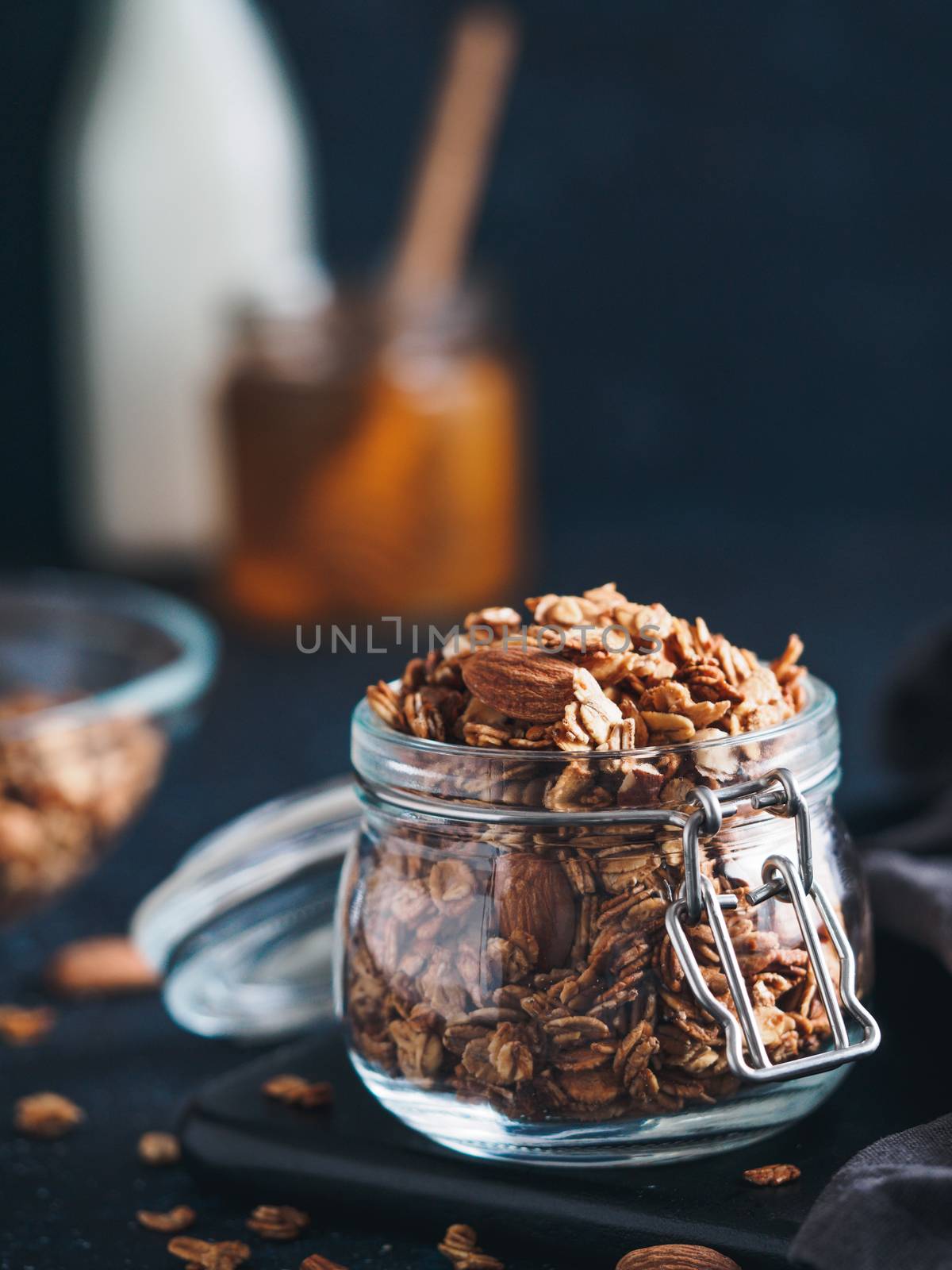 Homemade granola in glass jar on dark table. Ingredients for healthy breakfast - granola, milk and honey. Copy space for text. Low key.