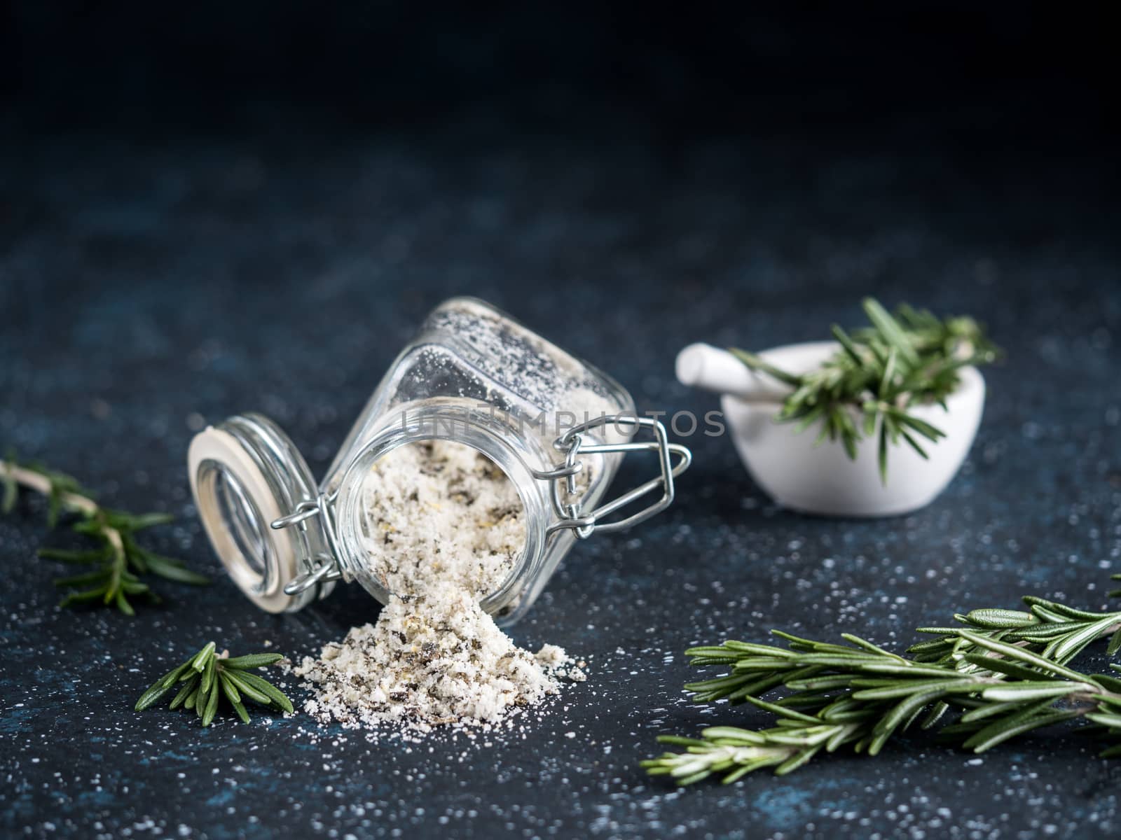 Sea salt scented herb rosemary and lemon zest. Sea salt with aromatic herb spilled out of glass jar on dark blue background. Scented salt and ingerdients. Copy space