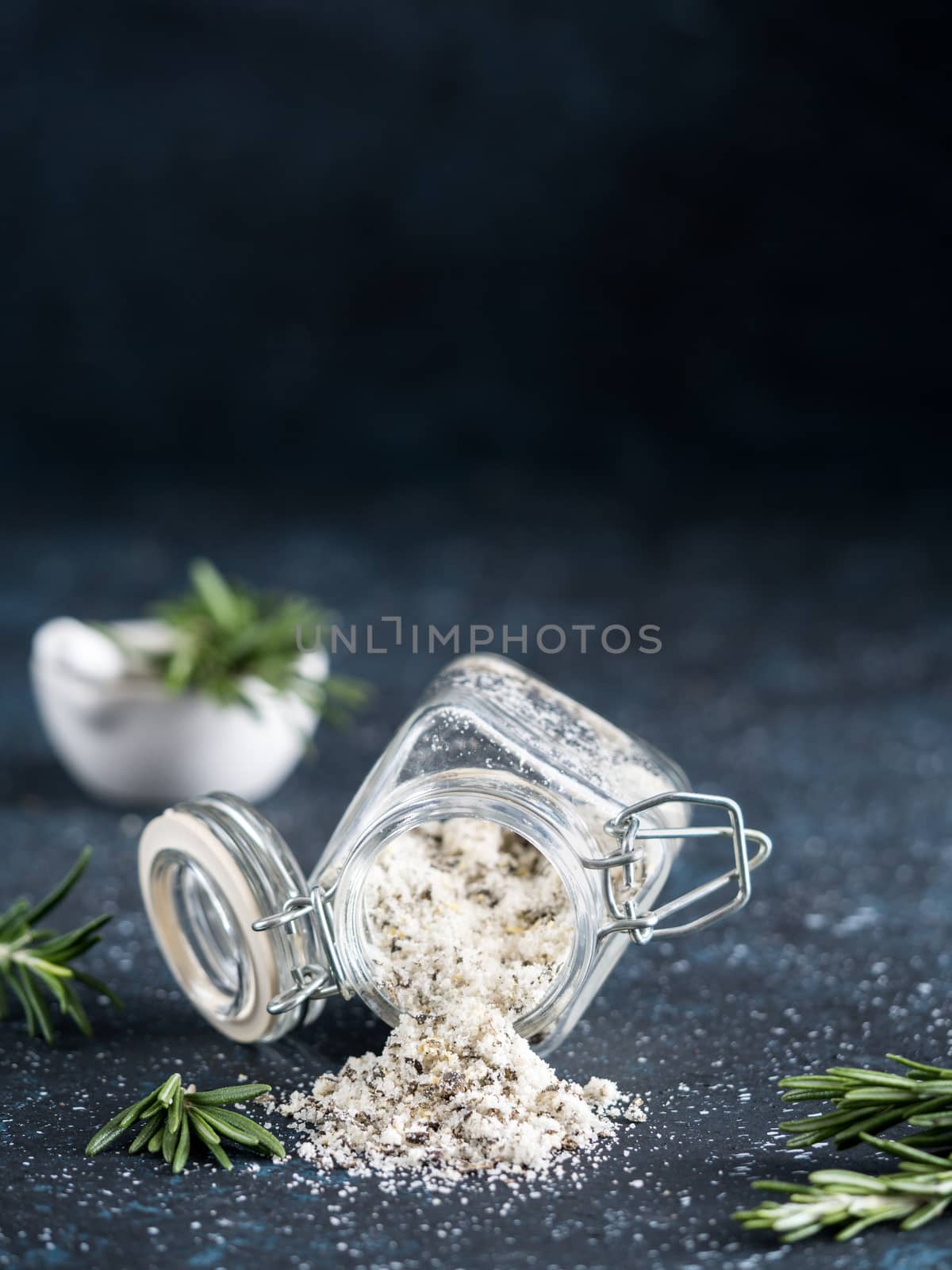 Sea salt scented herb rosemary and lemon zest. Sea salt with aromatic herb spilled out of glass jar on dark blue background. Scented salt and ingerdients. Copy space