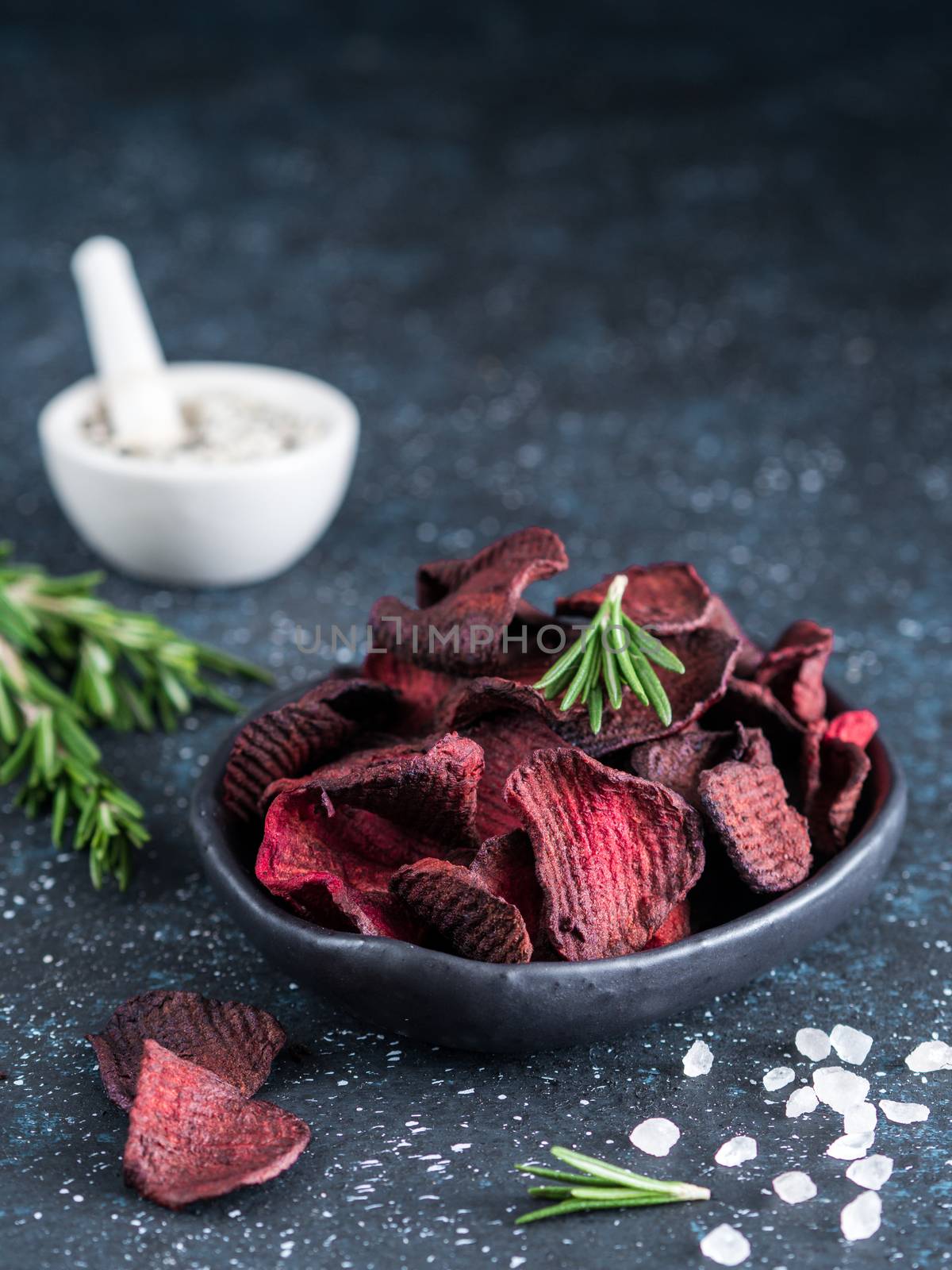 Baked beet slices with coarse sea salt scented rosemary. Vegan diet food idea and recipe. Healthy homemade beetroot chips in plate and ingredients on dark blue background. Copy space