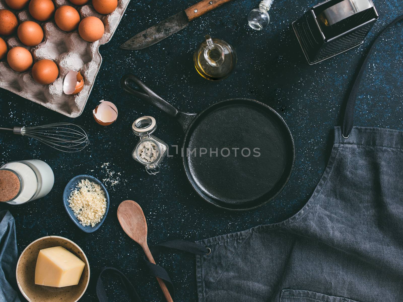 Empty cast-iron frying pan and ingredients for omlette or scrambled eggs,top view.Egg,shell,milk,parmesan cheese,scented rosemary salt,olive oil,pepper,apron,whisk,vintage knife ondark table.Copyspace