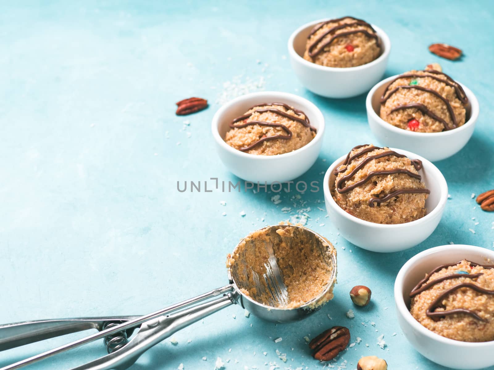 Safe-to-eat raw monster cookie dough in small portion bowl, ice cream scoop and nuts on blue background. Ideas and recipes for kids and toddlers meal. Copy space for text.