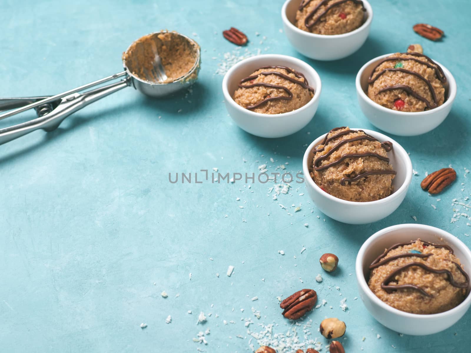 Safe-to-eat raw monster cookie dough in small portion bowl, ice cream scoop and nuts on blue background. Ideas and recipes for kids and toddlers meal. Copy space for text.