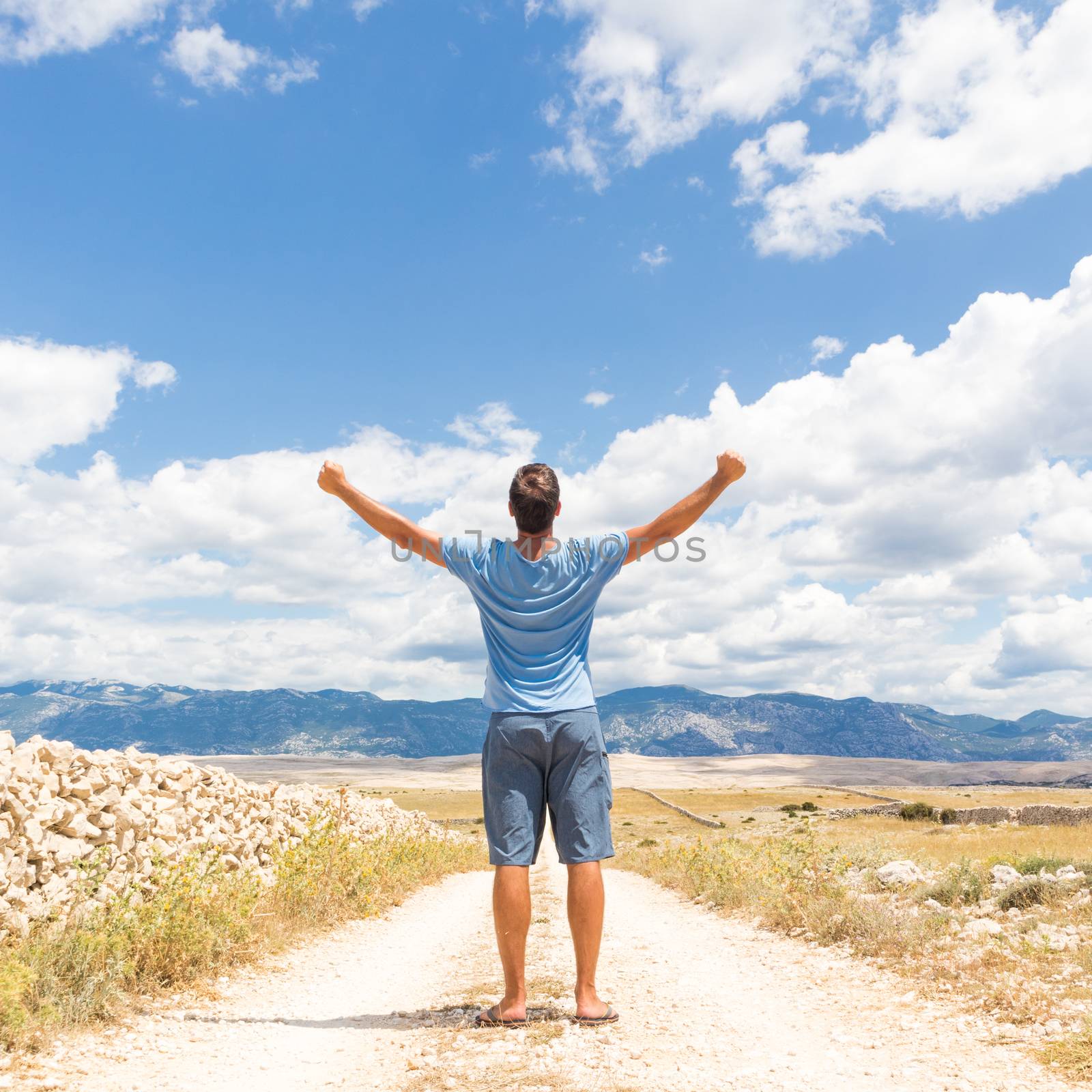 Rear view of casual sporty man standing on a dirt country road rising hands up to the clouds on a blue summer sky. Freedom and travel adventure concept. by kasto