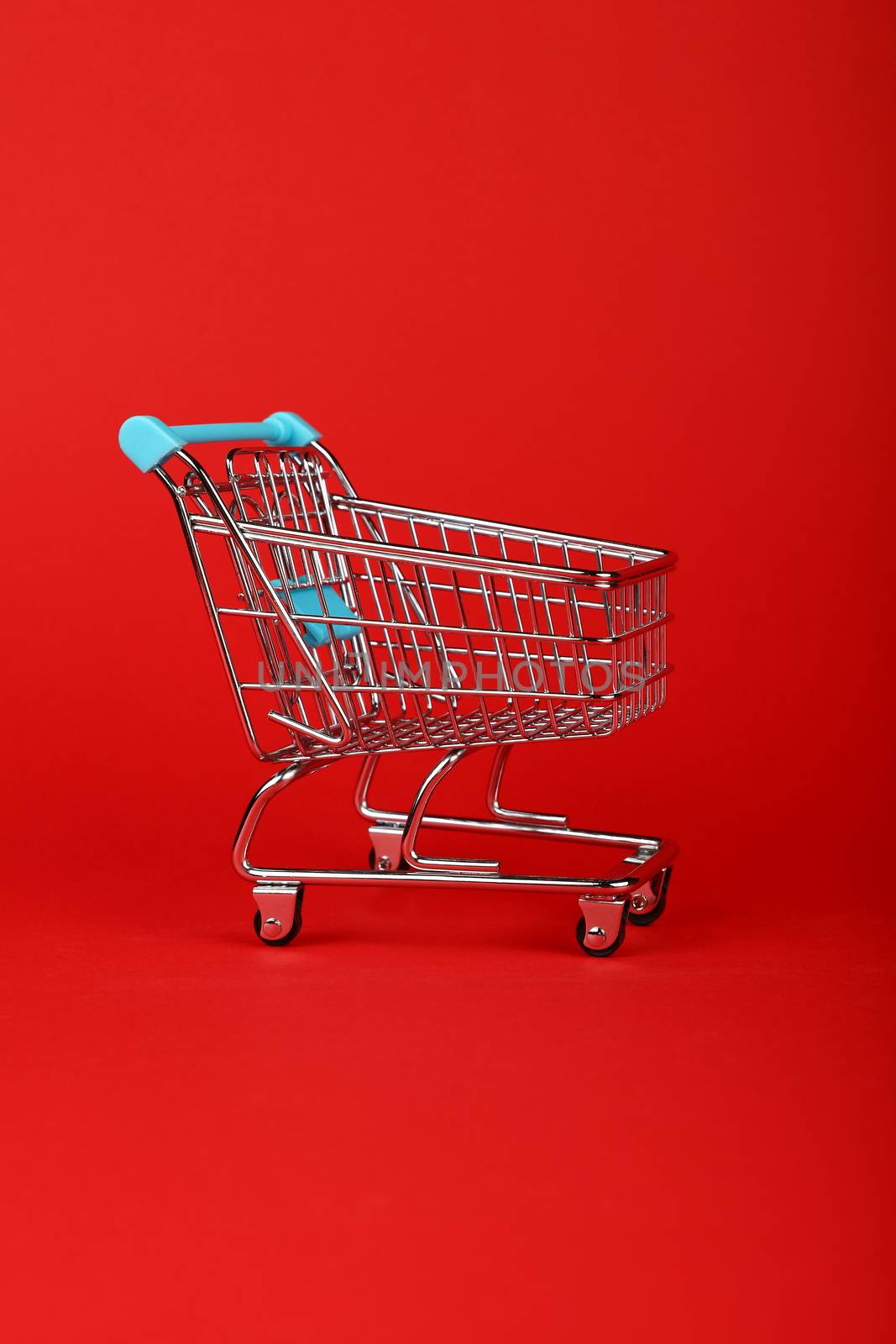 Close up empty toy metal supermarket shopping cart over vivid red background with copy space, low angle side view