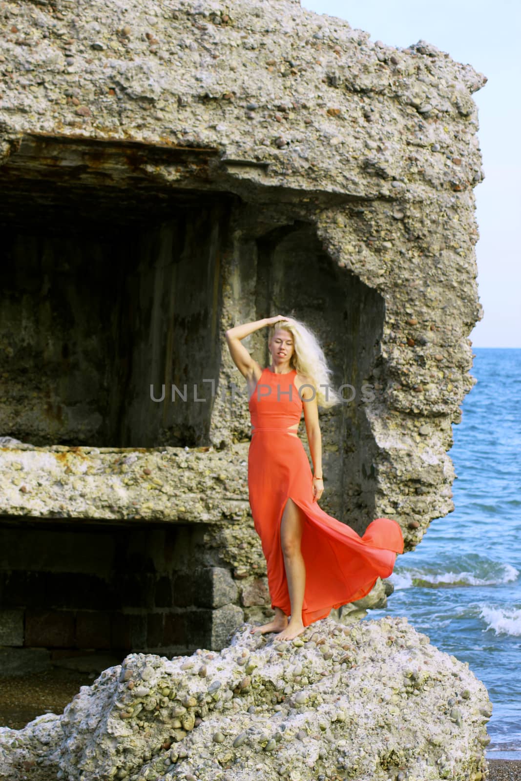 Tanned pretty blonde girl in a red dress stands on the stone ruins of an old fort, against the backdrop of the waves of the Baltic Sea.