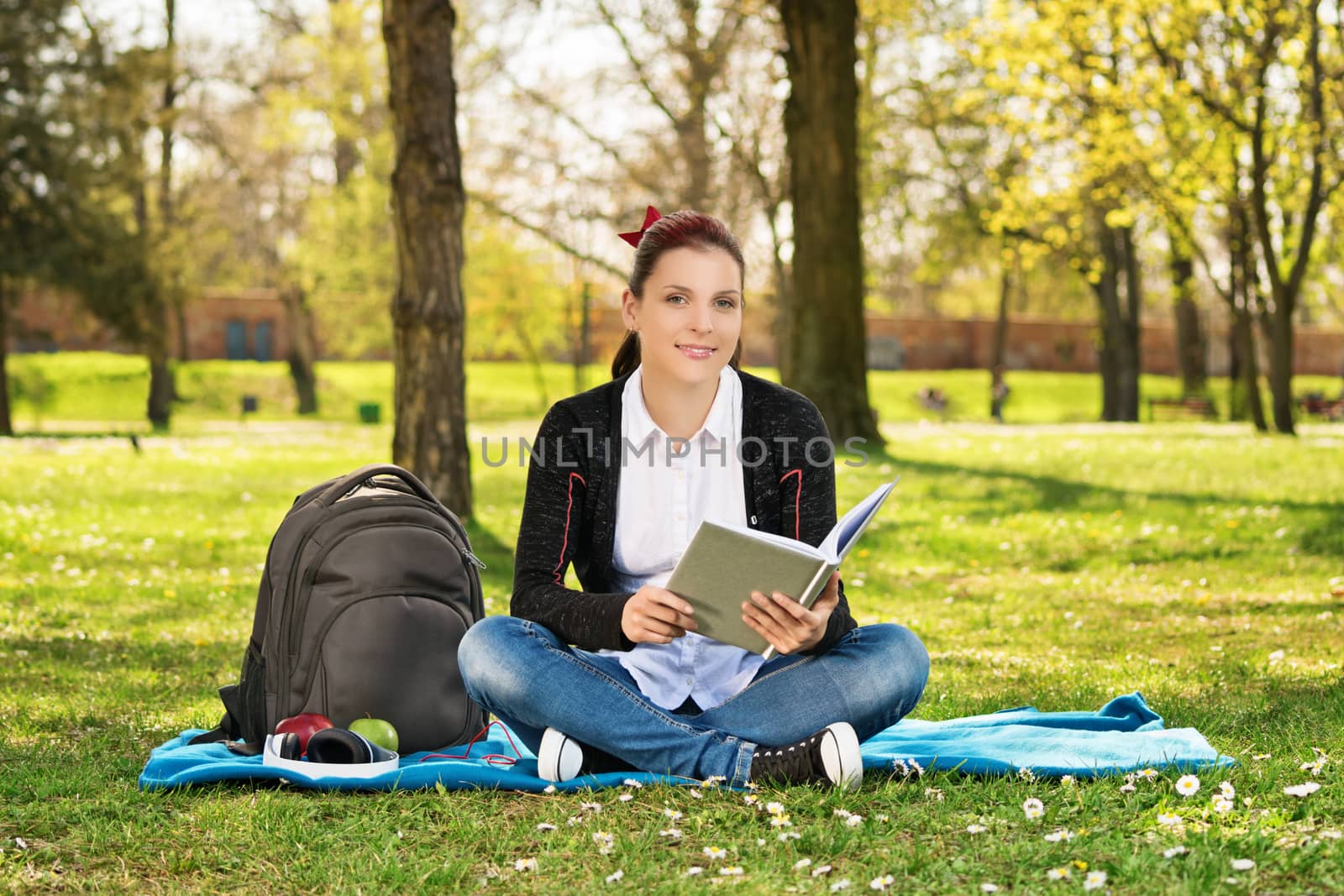 A portrait of a beautiful young female student holding a book, sitting on the grass in a park.