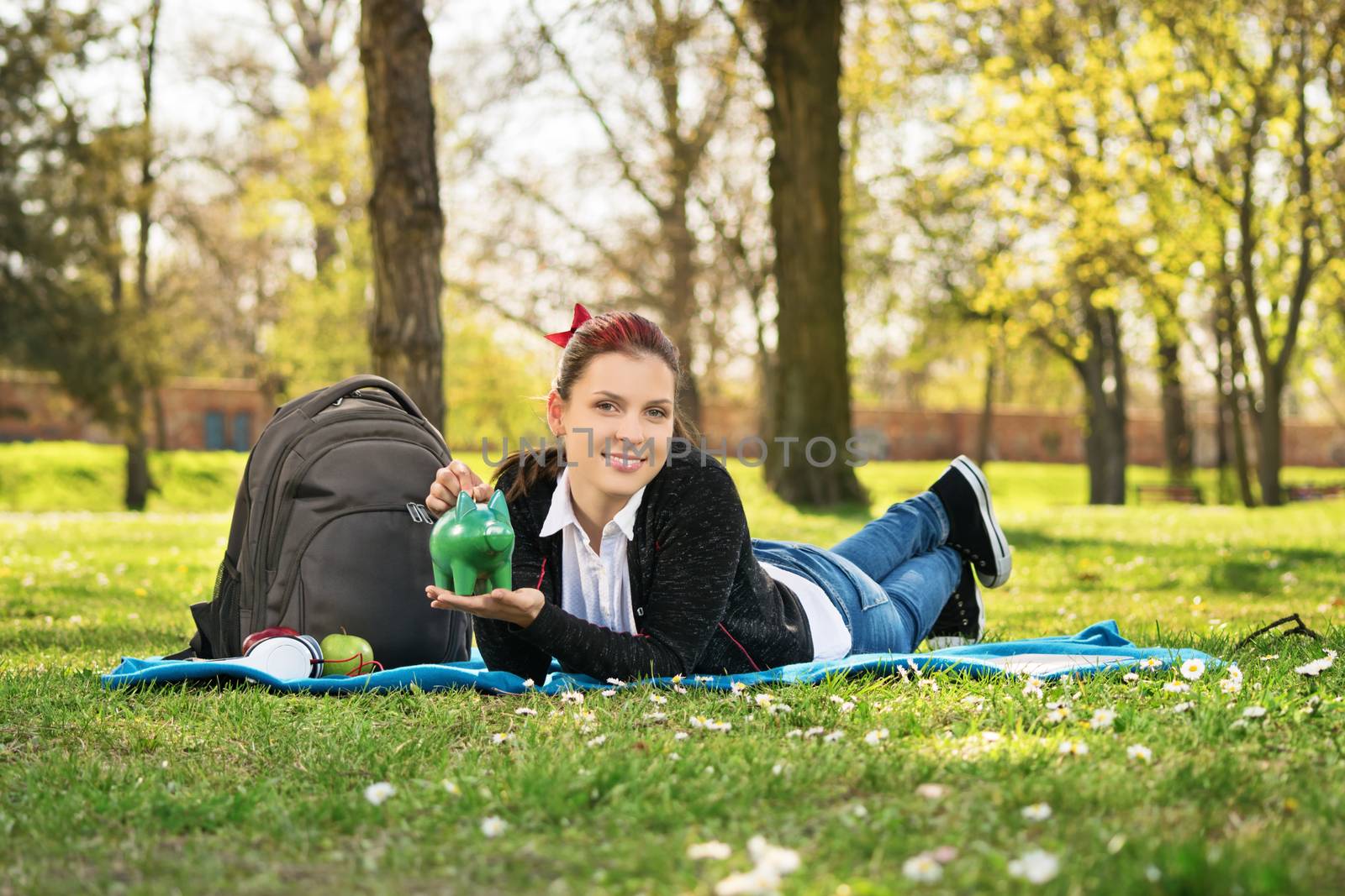 Thinking of my future and saving with ease. Beautiful young female student lying on a green grass in the park, holding a piggy bank.