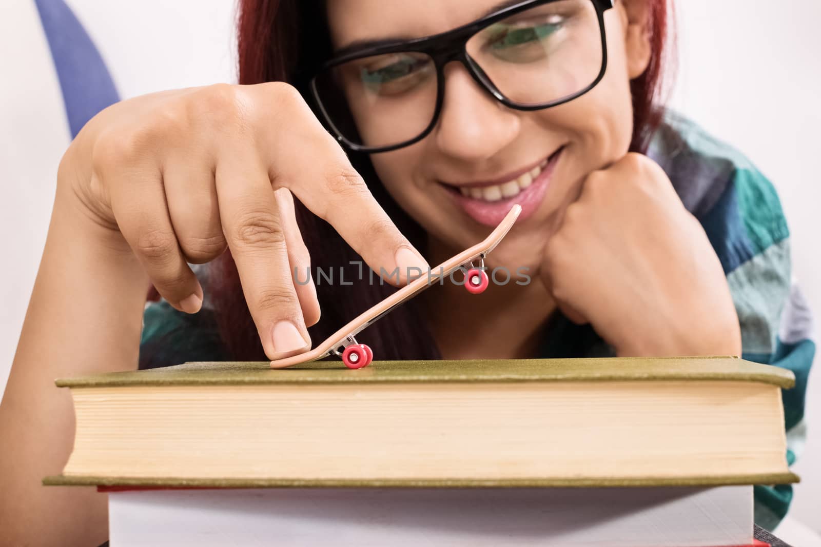 Close-up shot of a smiling young girl lying on a sofa, playing with toy skateboard on a stack of books.