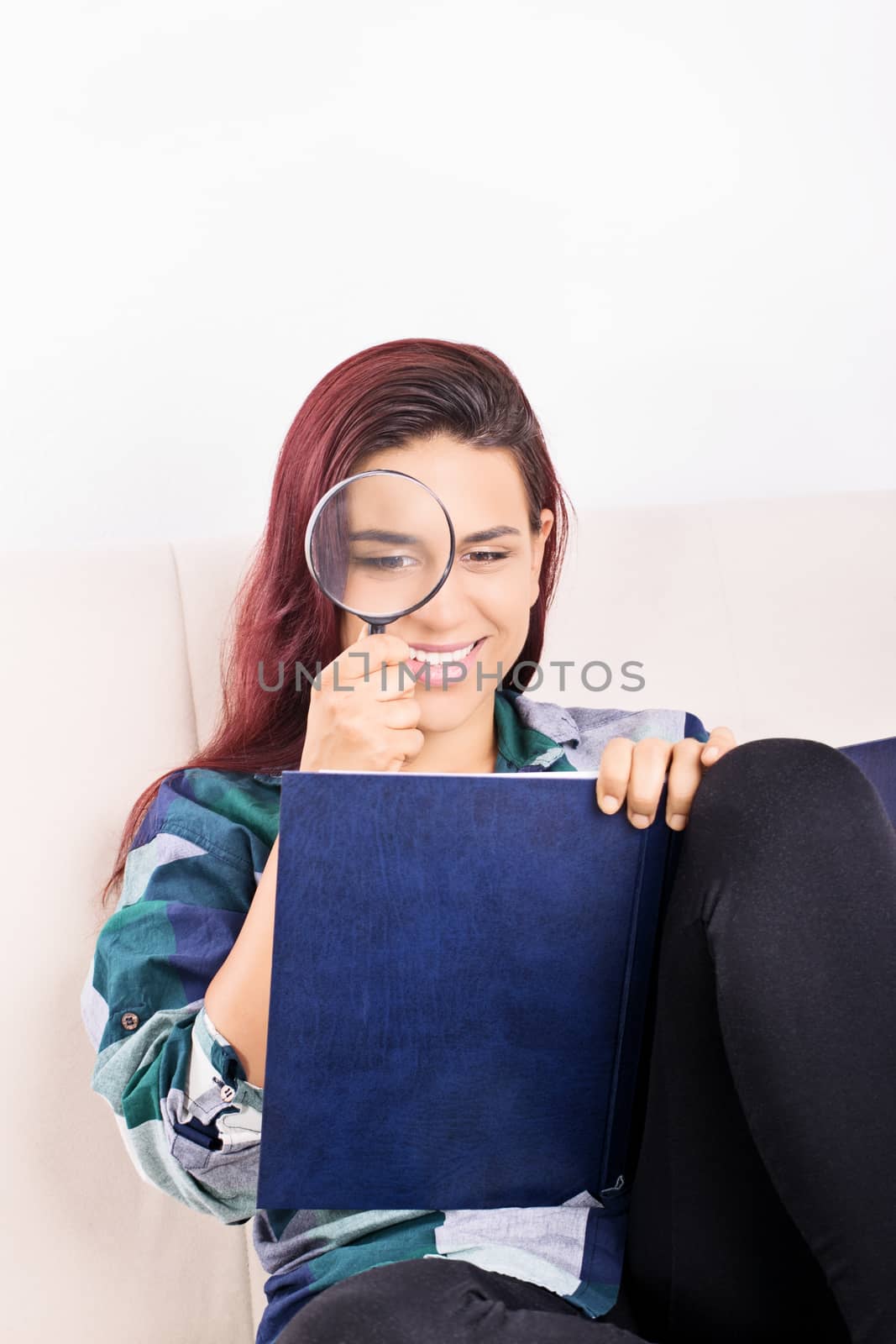 Beautiful young student girl, sitting on a couch looking at an open book through a magnifying glass.
