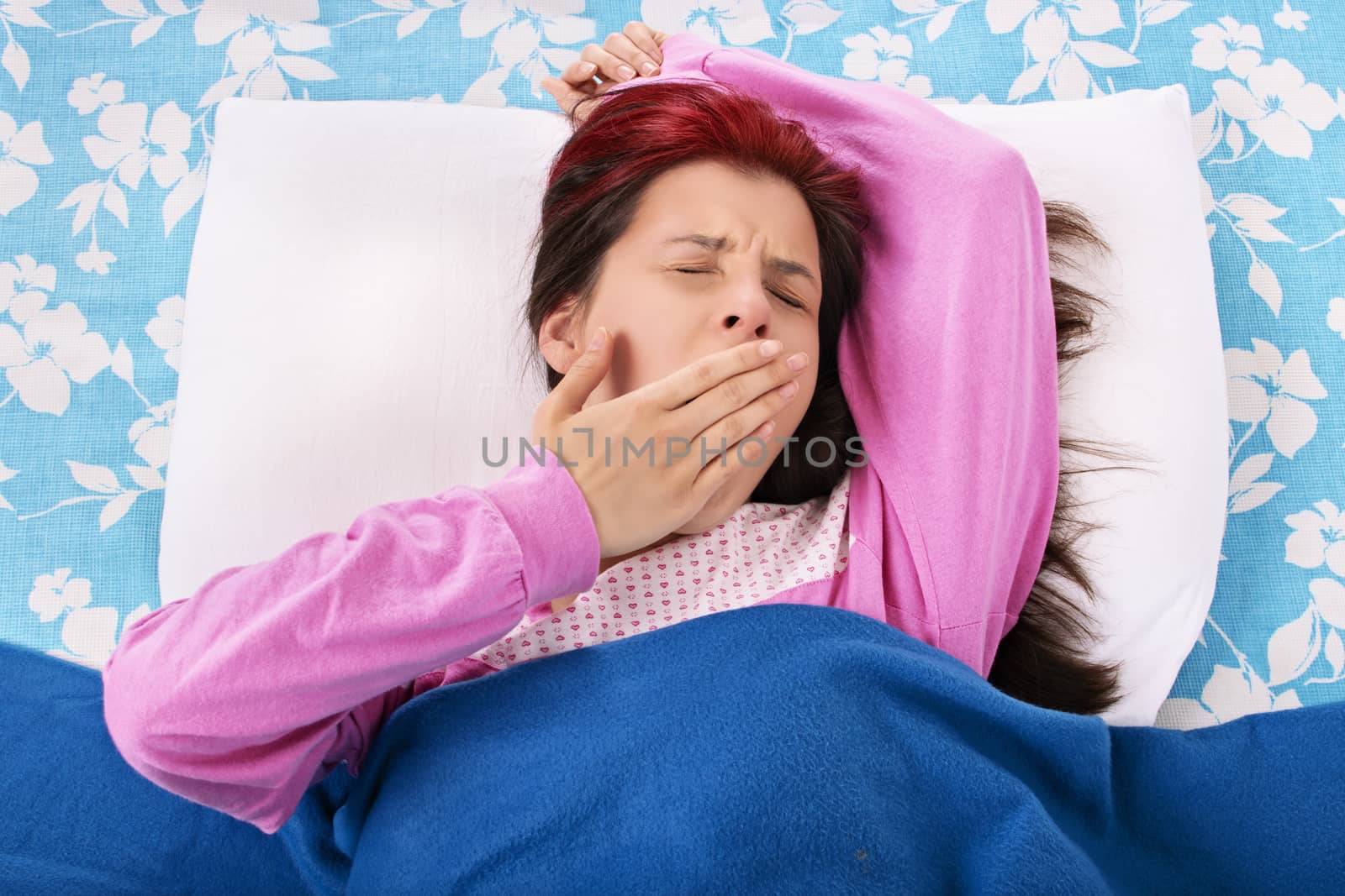 Young beautiful girl lying on a pillow under a blanket yawning, isolated on a white background.